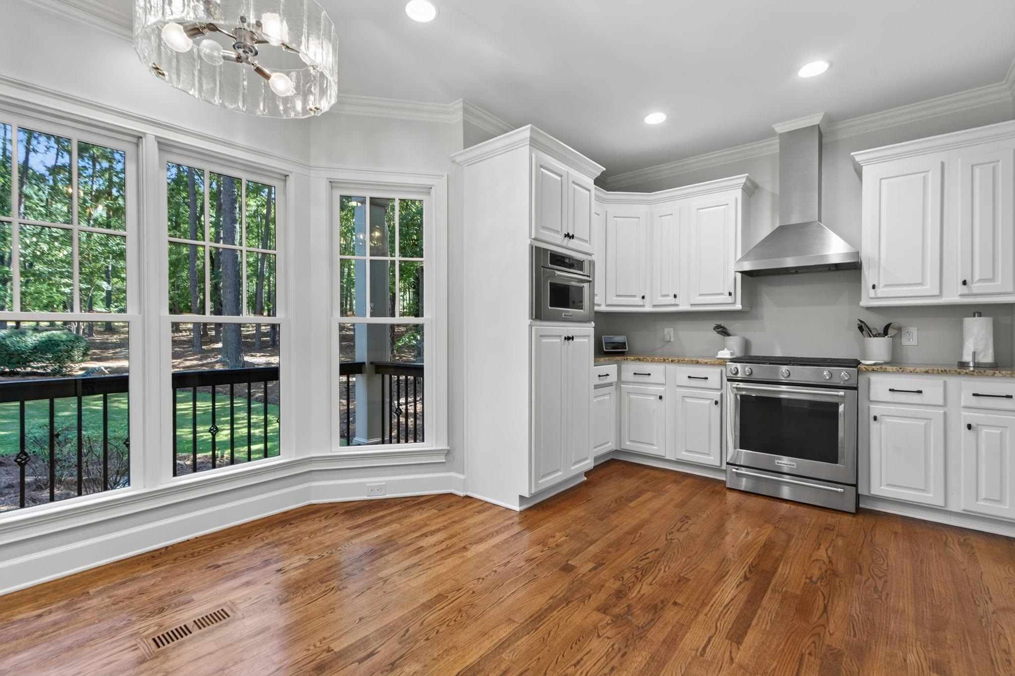 Kitchen with white cabinets, stainless steel appliances, hardwood floors, and tall windows overlooking trees.