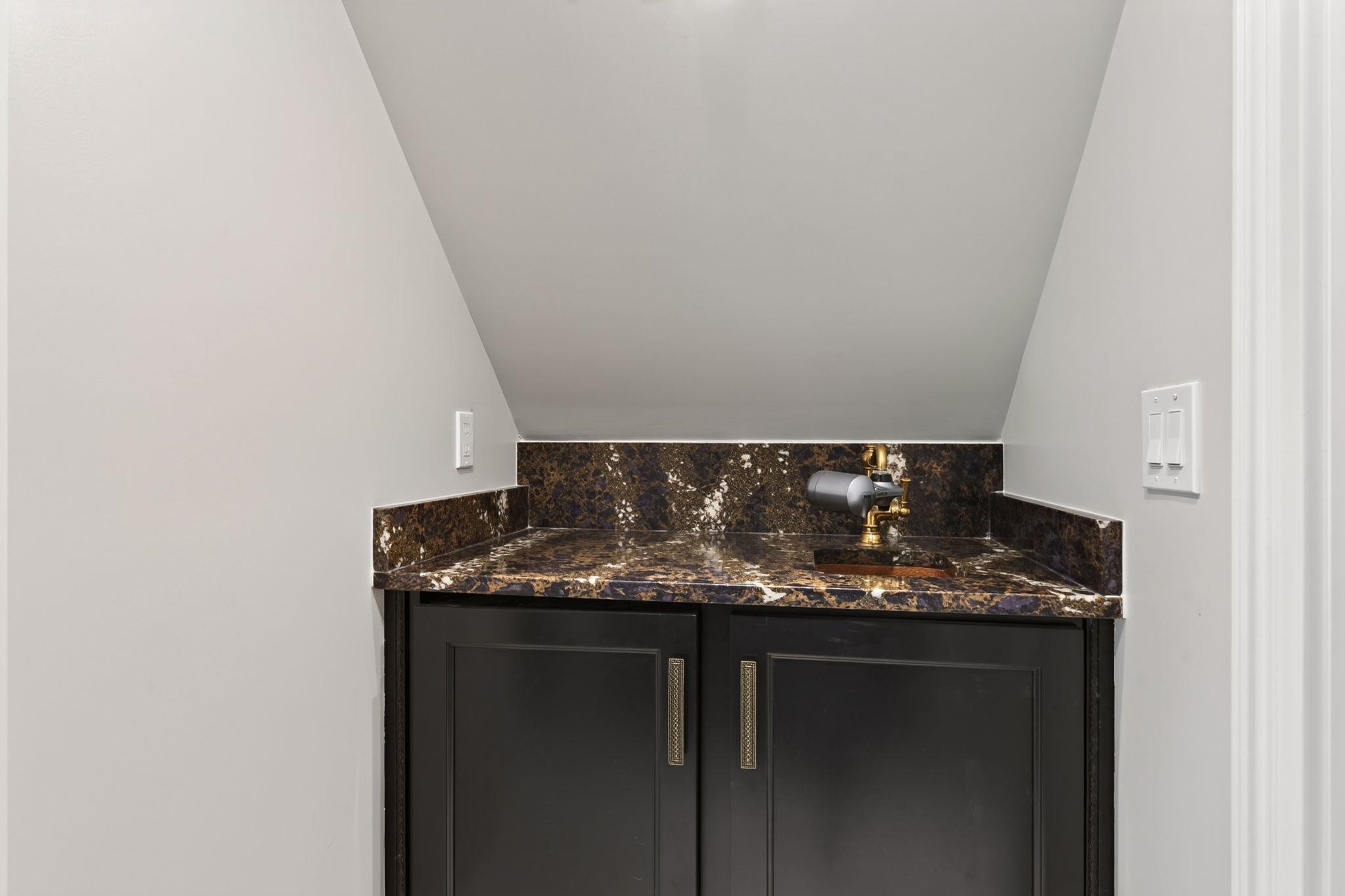 Bathroom vanity with dark cabinets, veined stone countertop, and brass faucet beneath a sloped ceiling.