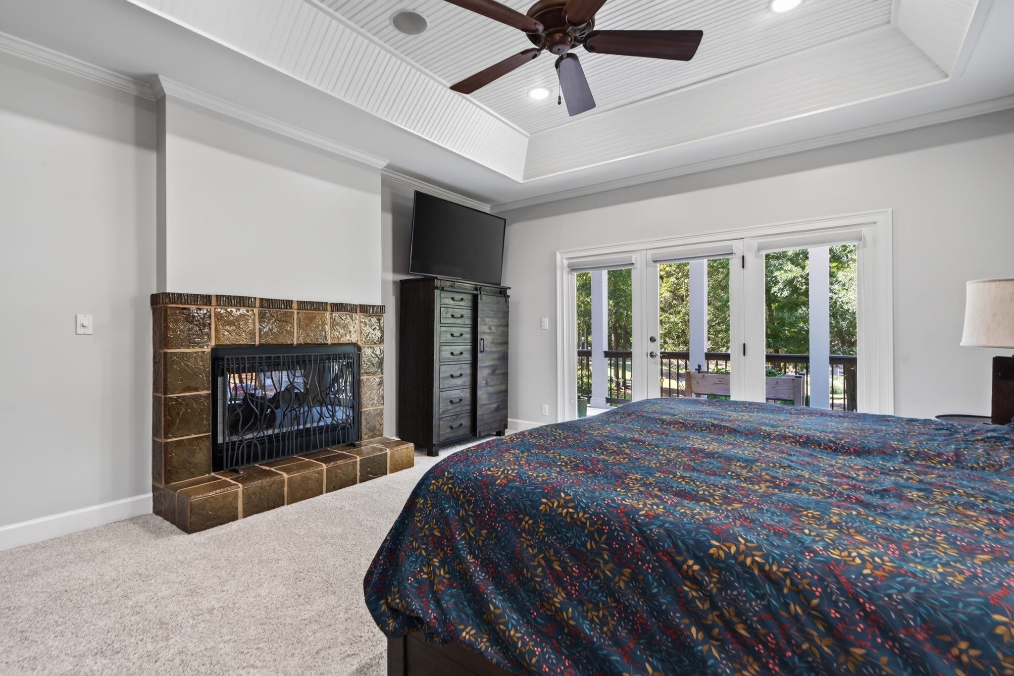 Bedroom with a tiled fireplace, dark ceiling fan, and glass doors to a wooded deck.