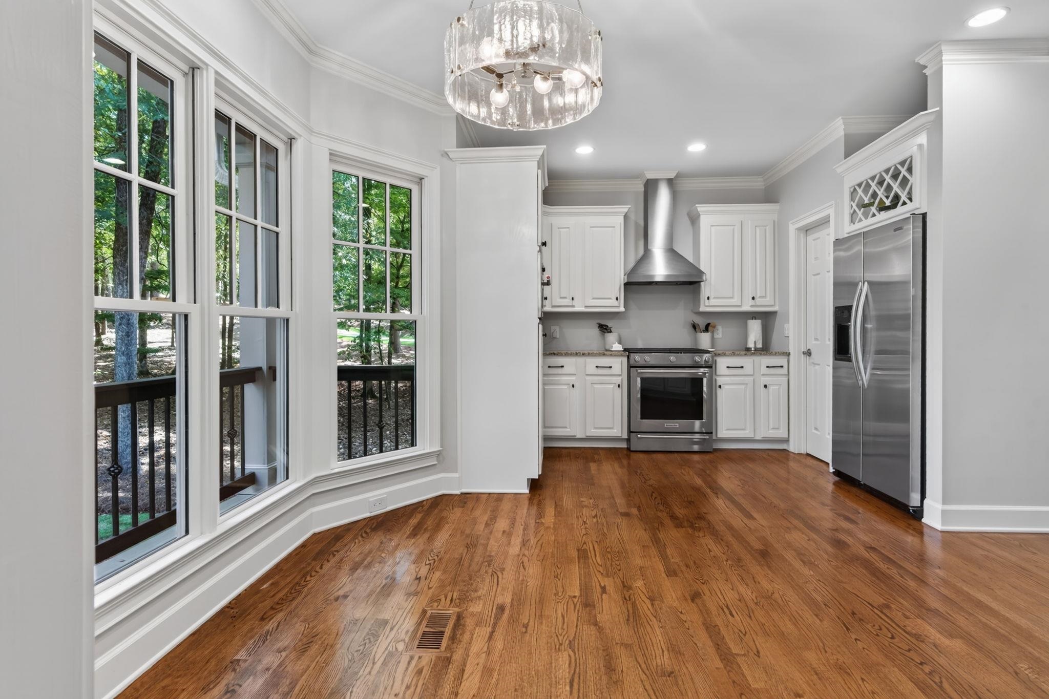 Bright white kitchen with stainless steel appliances, hardwood floors, and large windows facing trees.