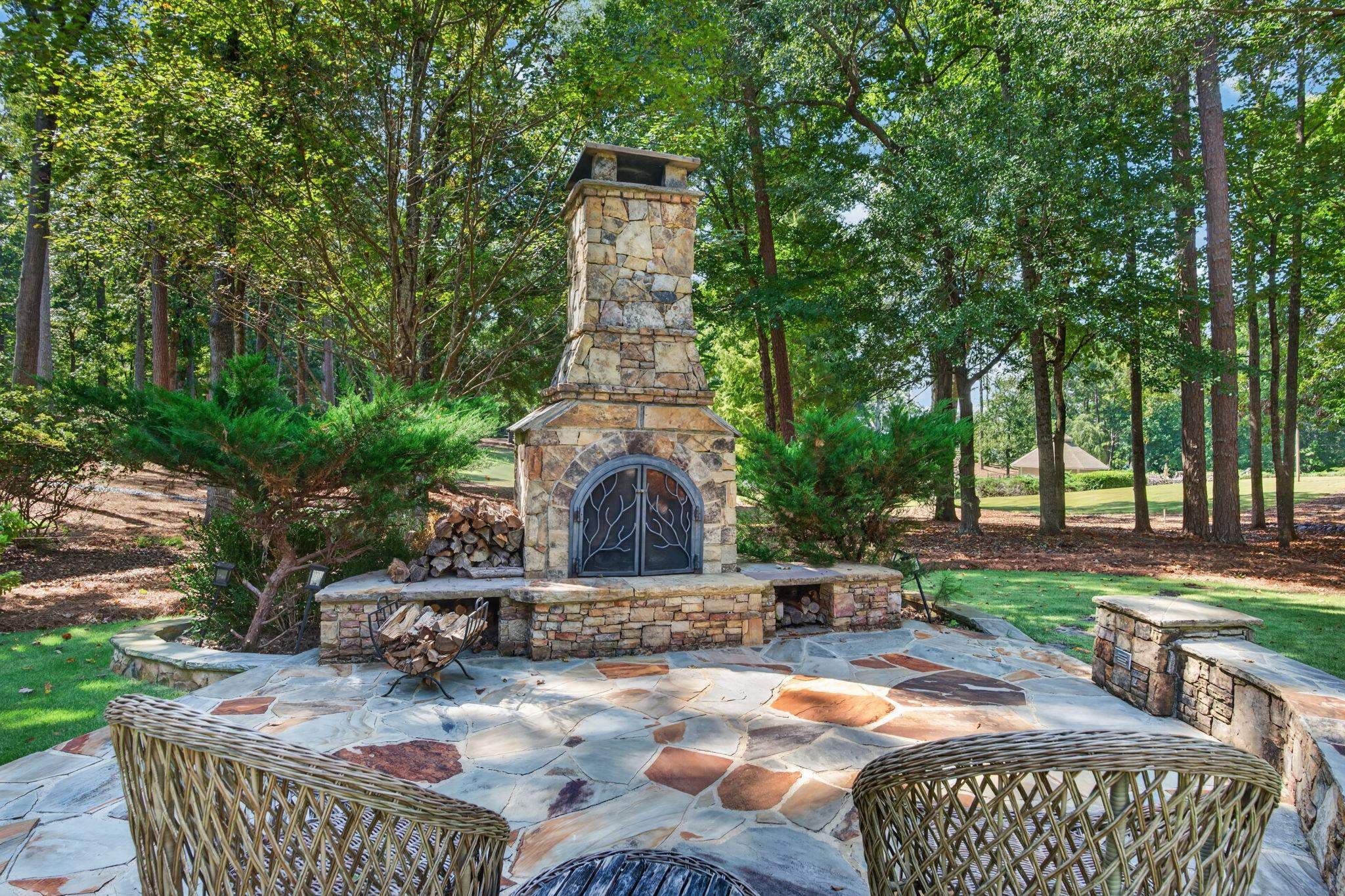 Stone outdoor fireplace on a flagstone patio, framed by trees and wicker chairs.