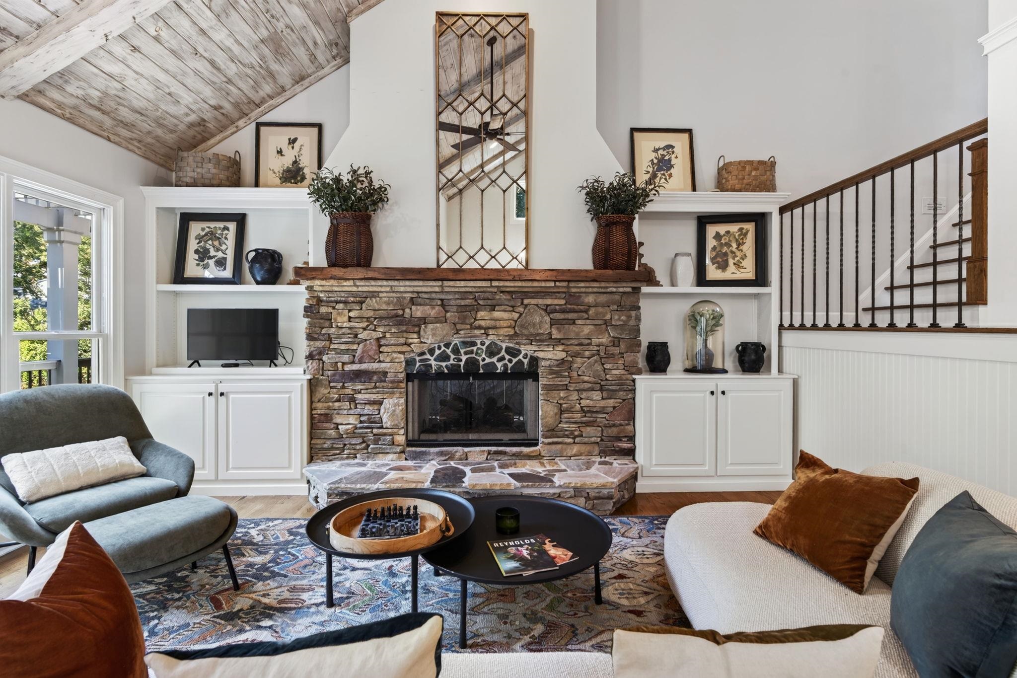 Living room with vaulted wood ceiling, stone fireplace, built-in shelves, and staircase.
