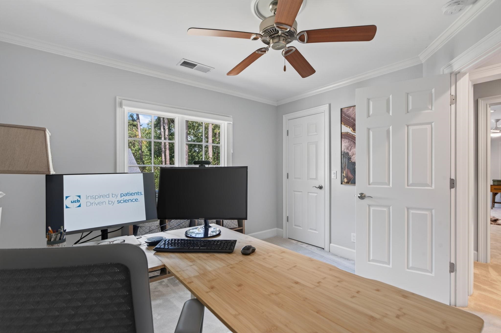 Home office with a long wooden desk, dual monitors, and windows overlooking trees.