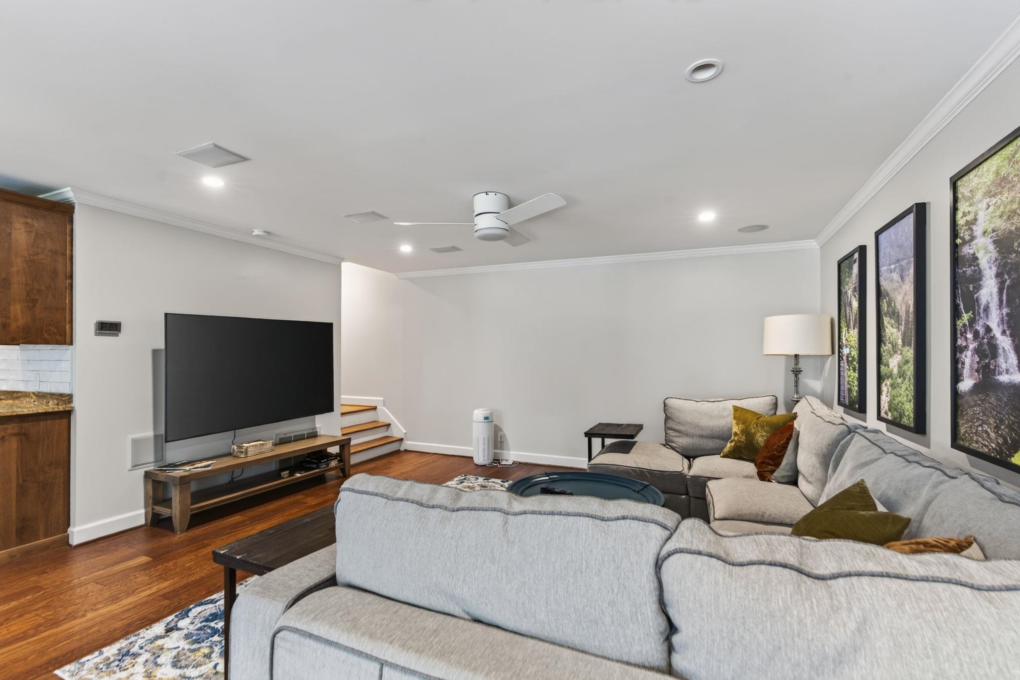 Living room with a large sectional sofa, wall-mounted TV, hardwood floors, and framed nature prints.