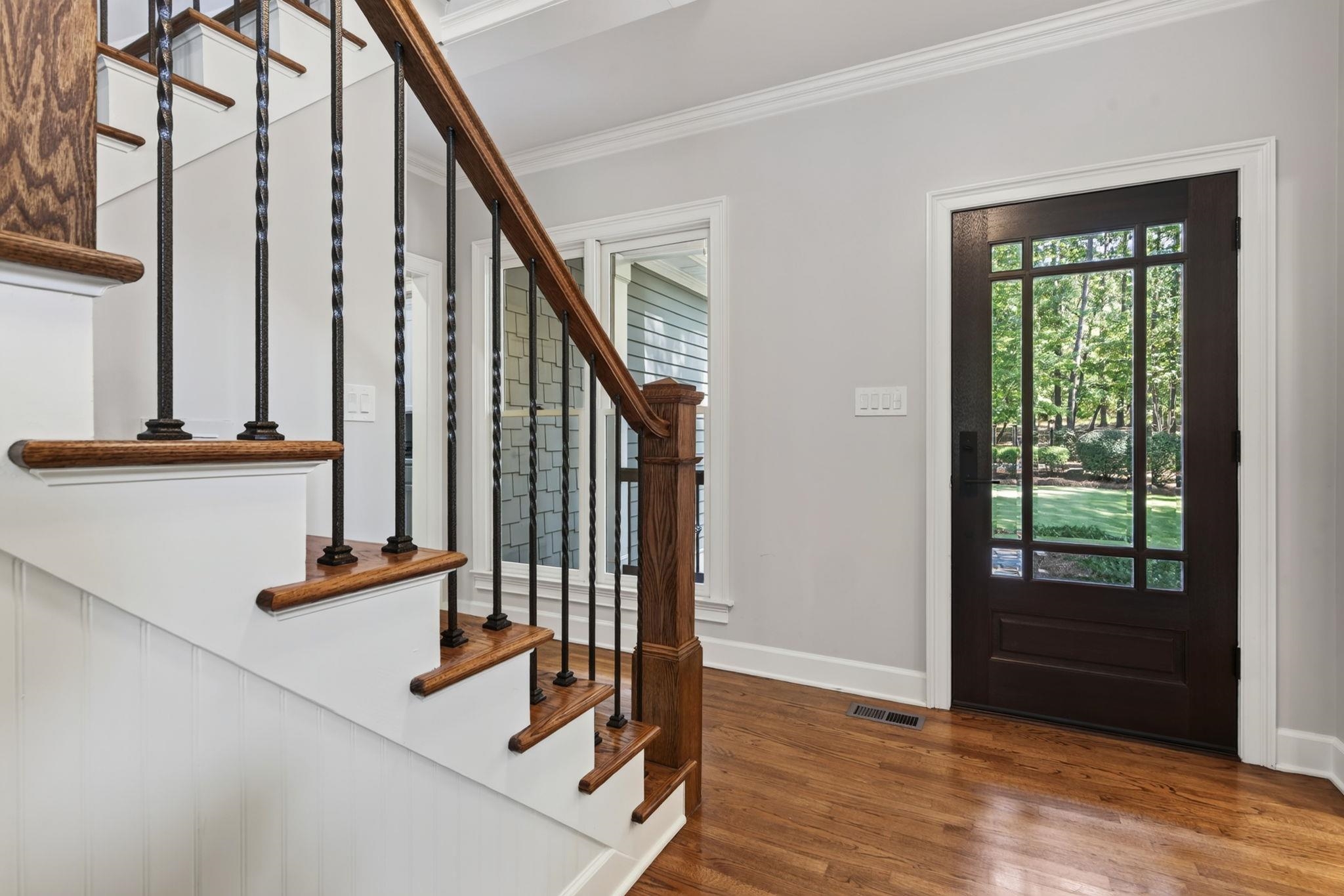 Entry foyer with a wood-and-metal staircase, hardwood floors, and a glass-paneled front door overlooking greenery.
