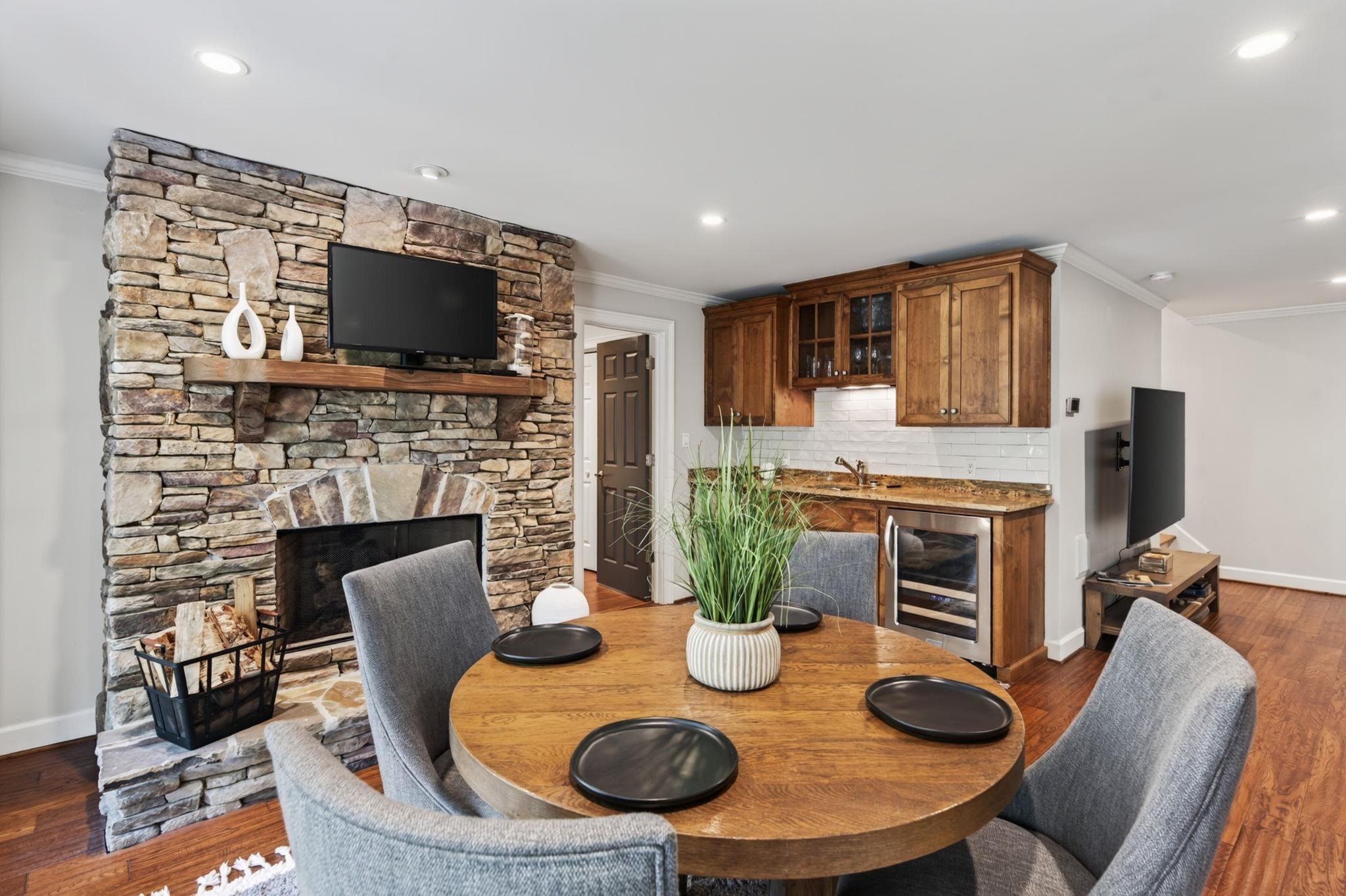 Dining area with round wooden table beside a stone fireplace and built-in wet bar.