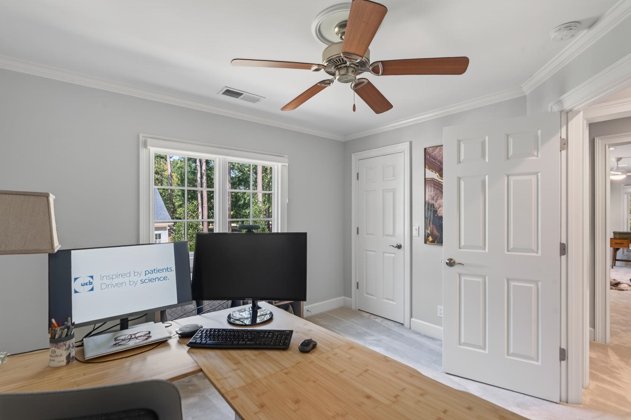 Home office with a wood desk and dual monitors beside windows overlooking trees.