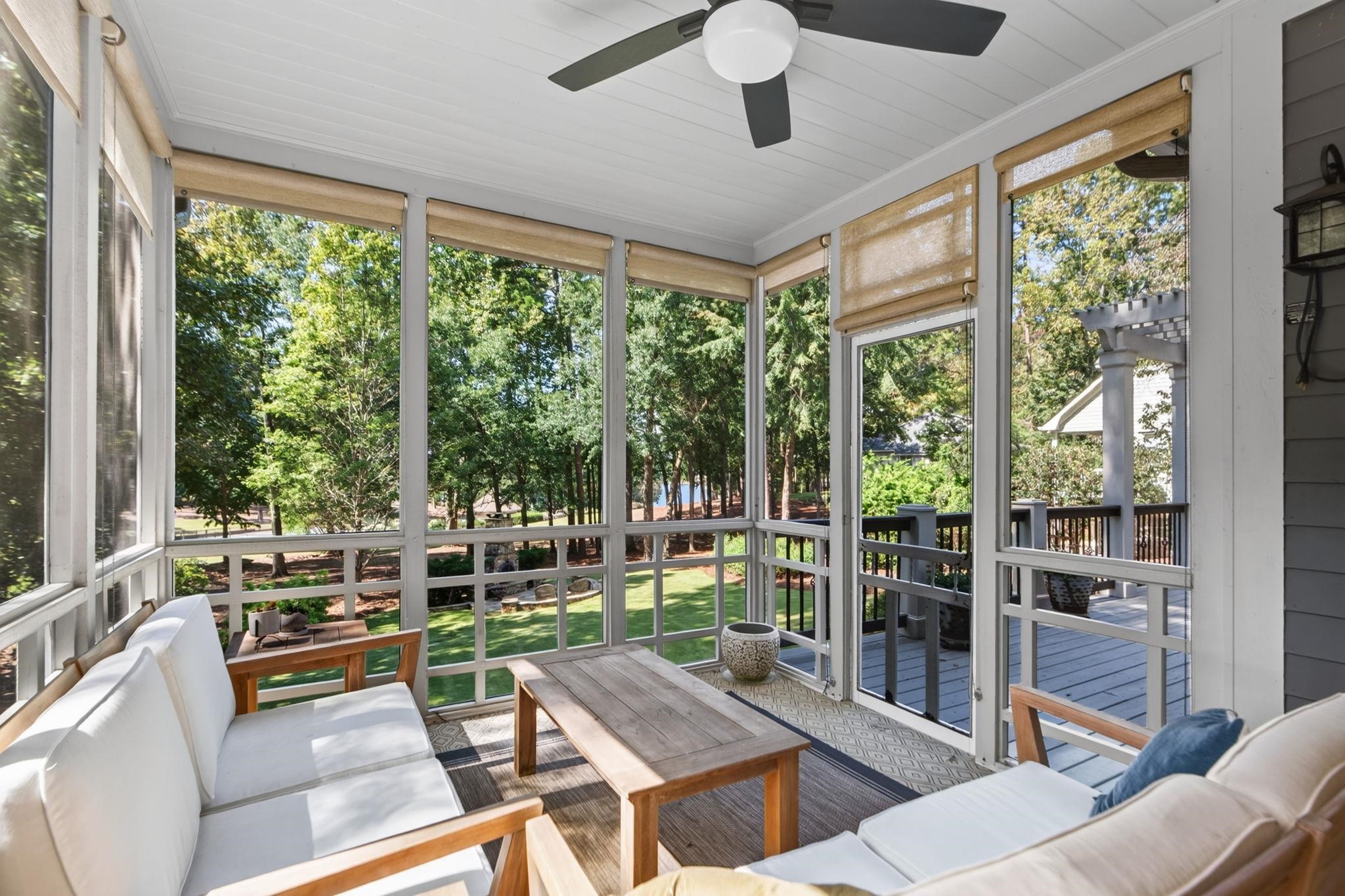 Furnished sunroom with screened windows overlooking a leafy yard and a glimpse of water.