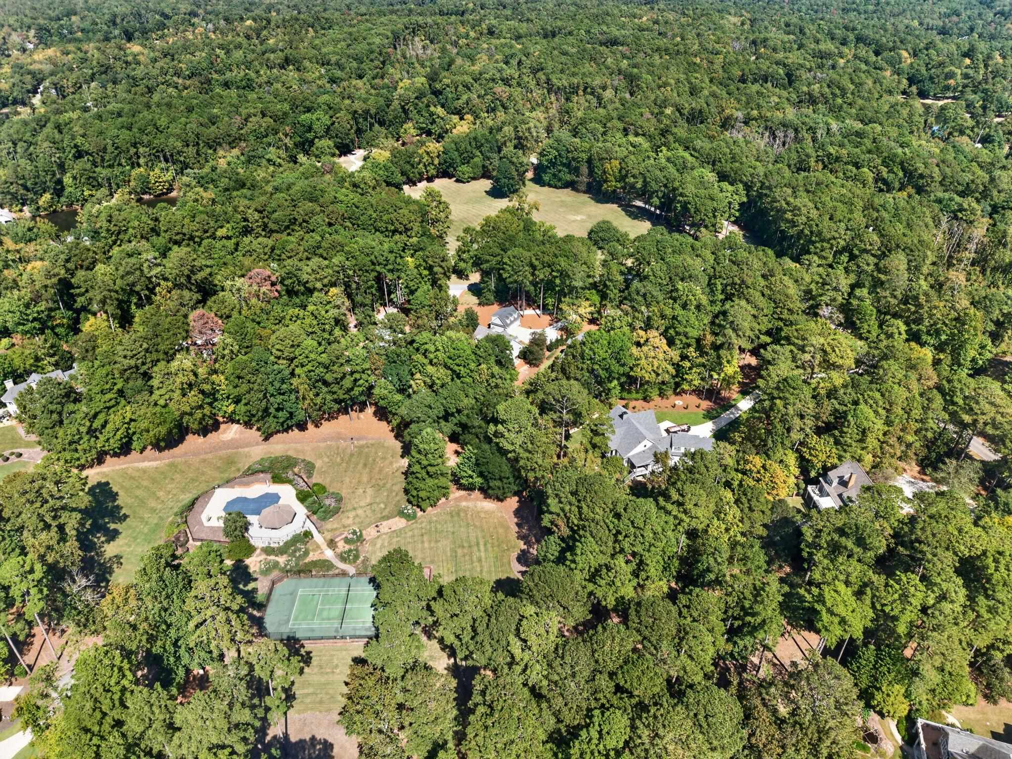 Aerial view of a wooded property with a pool, tennis court, and homes among landscaped lawns.