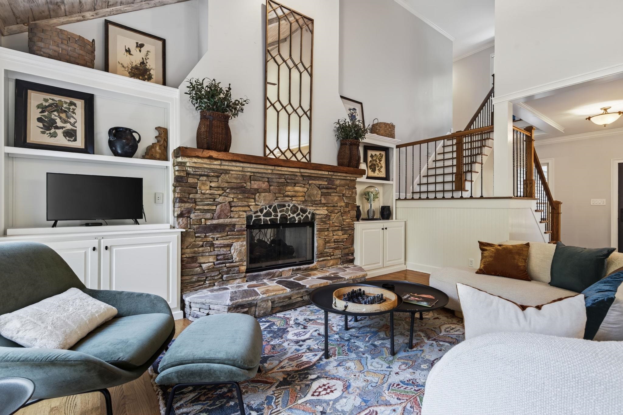 Living room with a stone fireplace, built-in cabinetry, exposed ceiling beams, and a staircase.