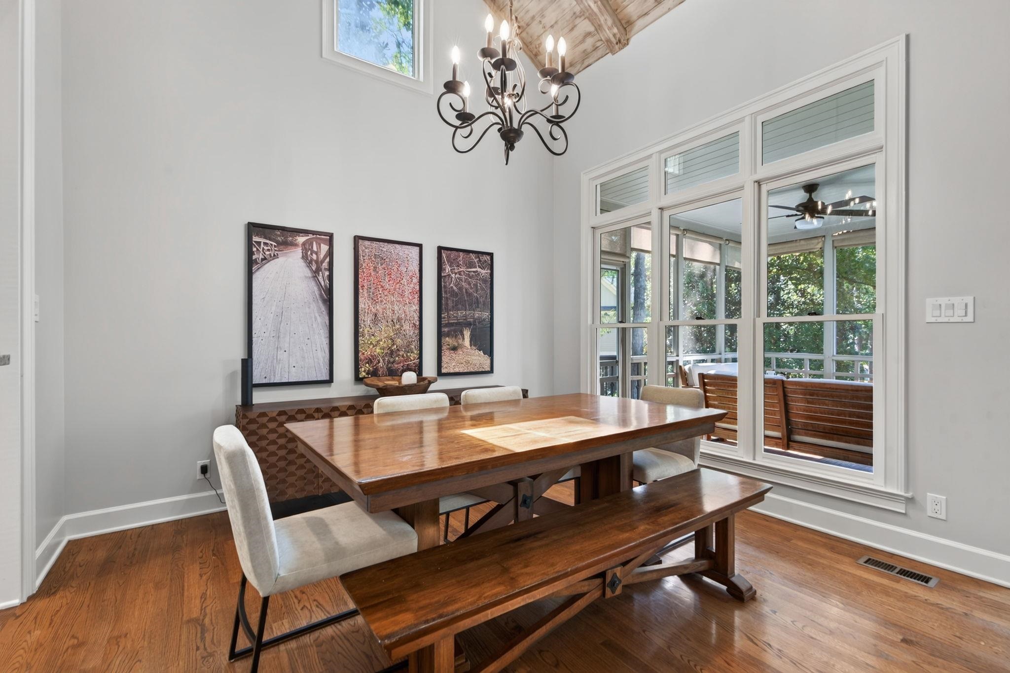 Dining room with a wooden table and bench beneath a chandelier, beside tall windows overlooking a screened porch.