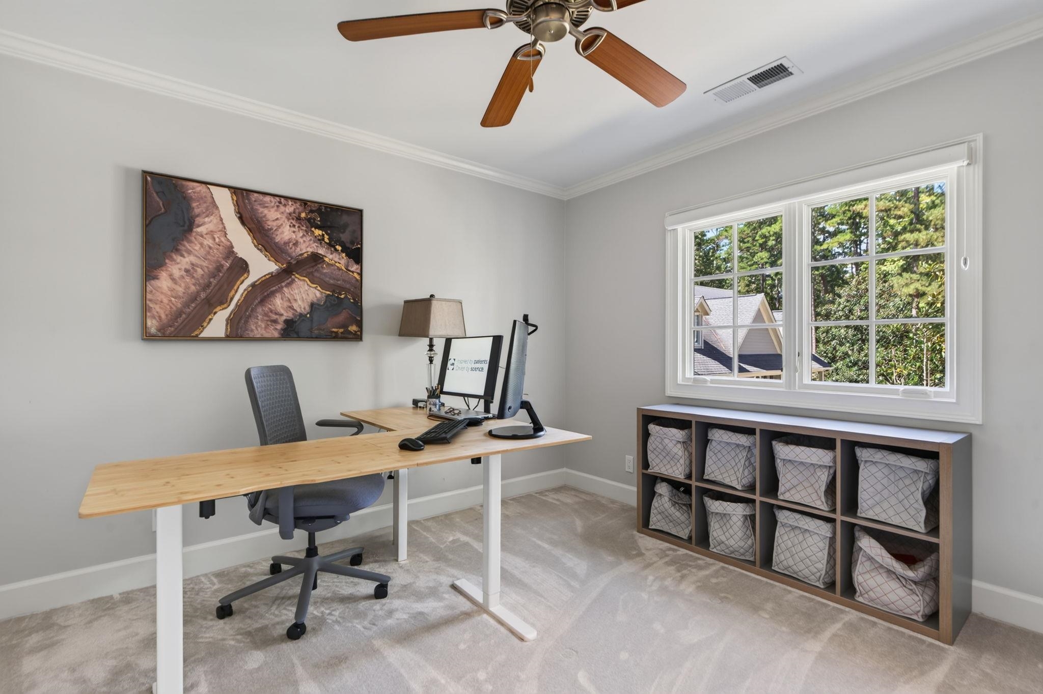 Home office with a wooden standing desk, gray chair, and cubby storage beneath a large window.