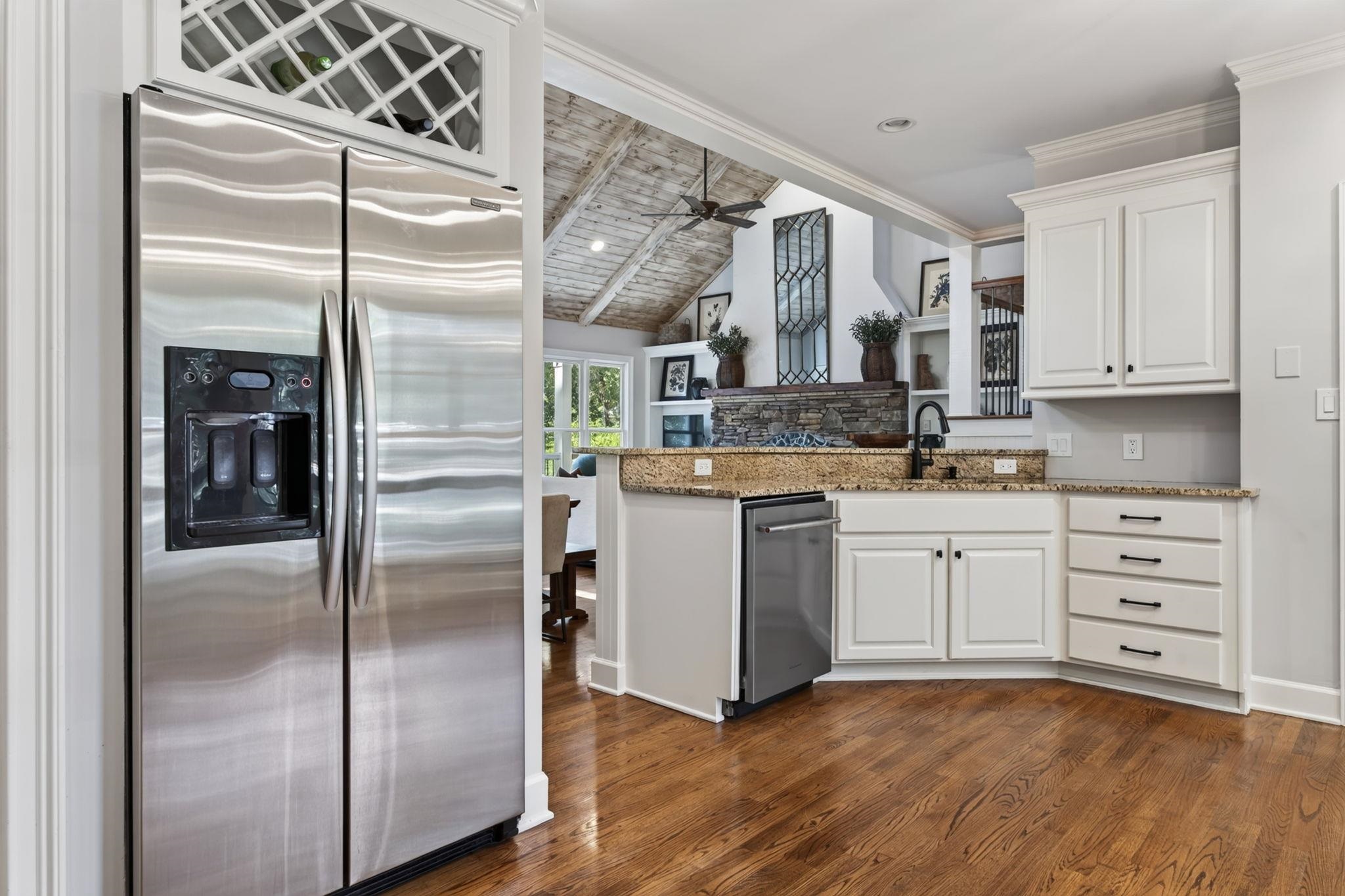 Kitchen with white cabinets, granite counters, stainless appliances, and a view of a stone fireplace beneath a vaulted ceiling.