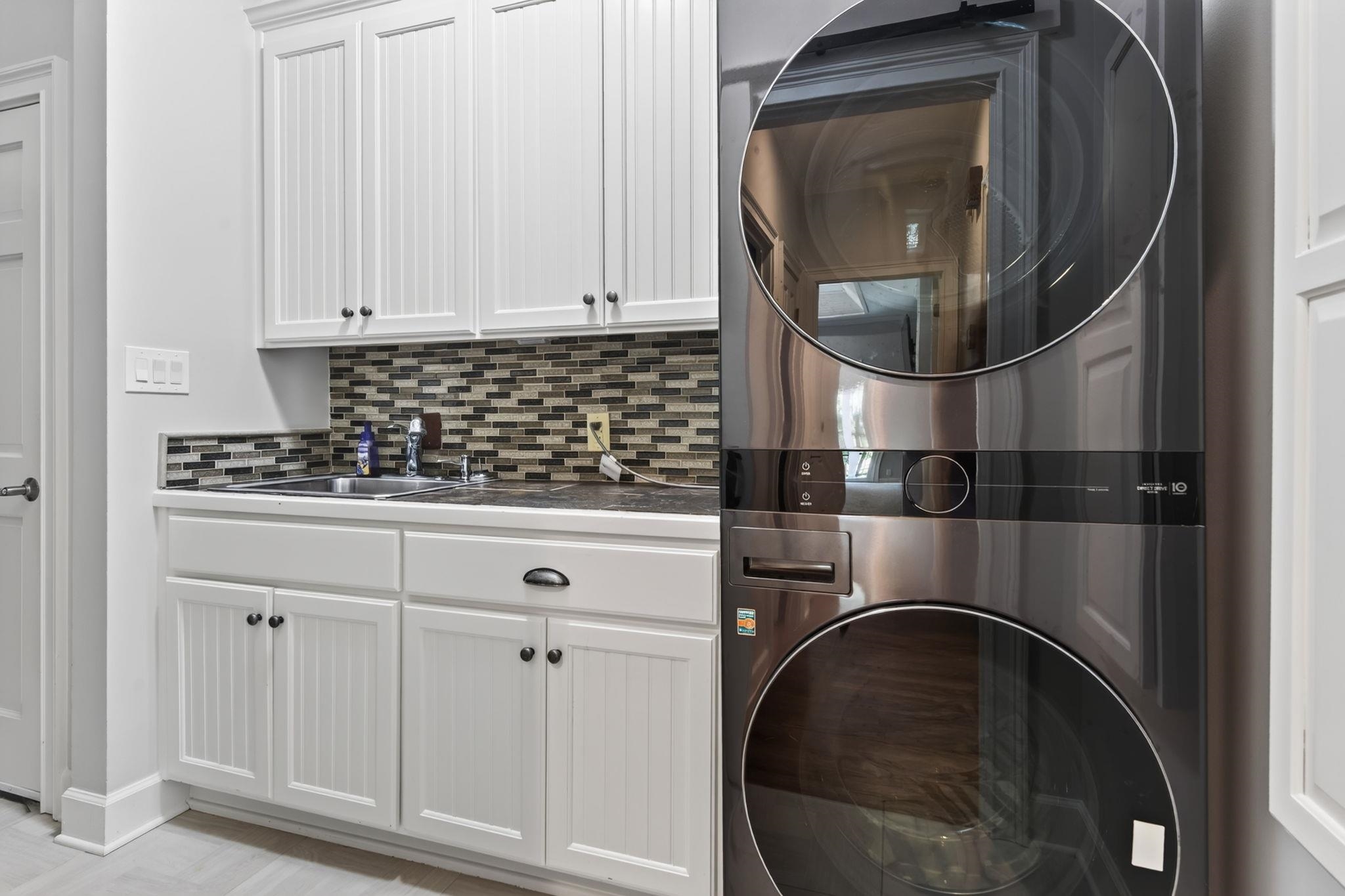 Laundry room with white cabinets, a sink, mosaic tile backsplash, and stacked stainless steel washer and dryer.