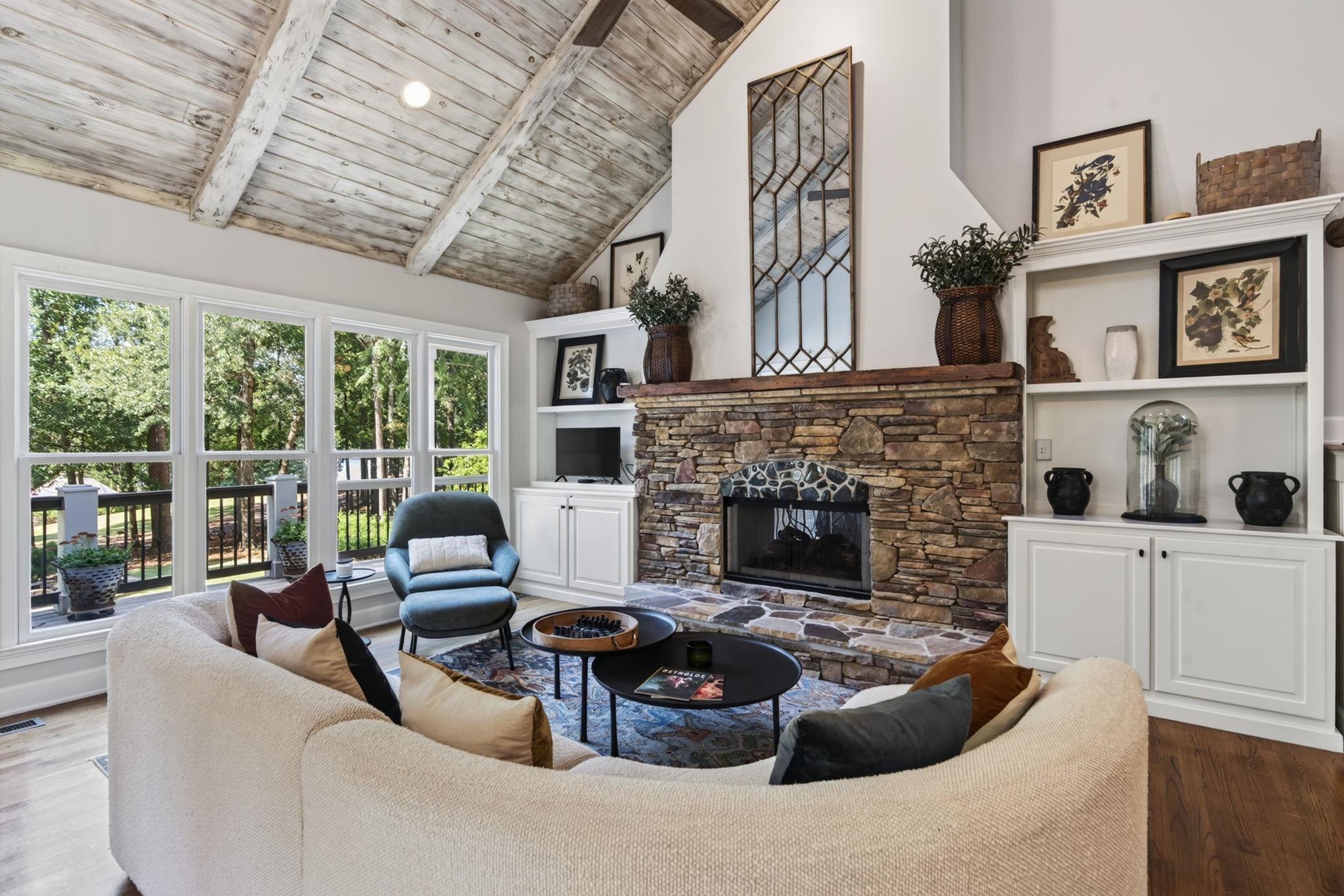 Living room with a vaulted wood ceiling, stone fireplace, built-ins, and tall windows overlooking trees.