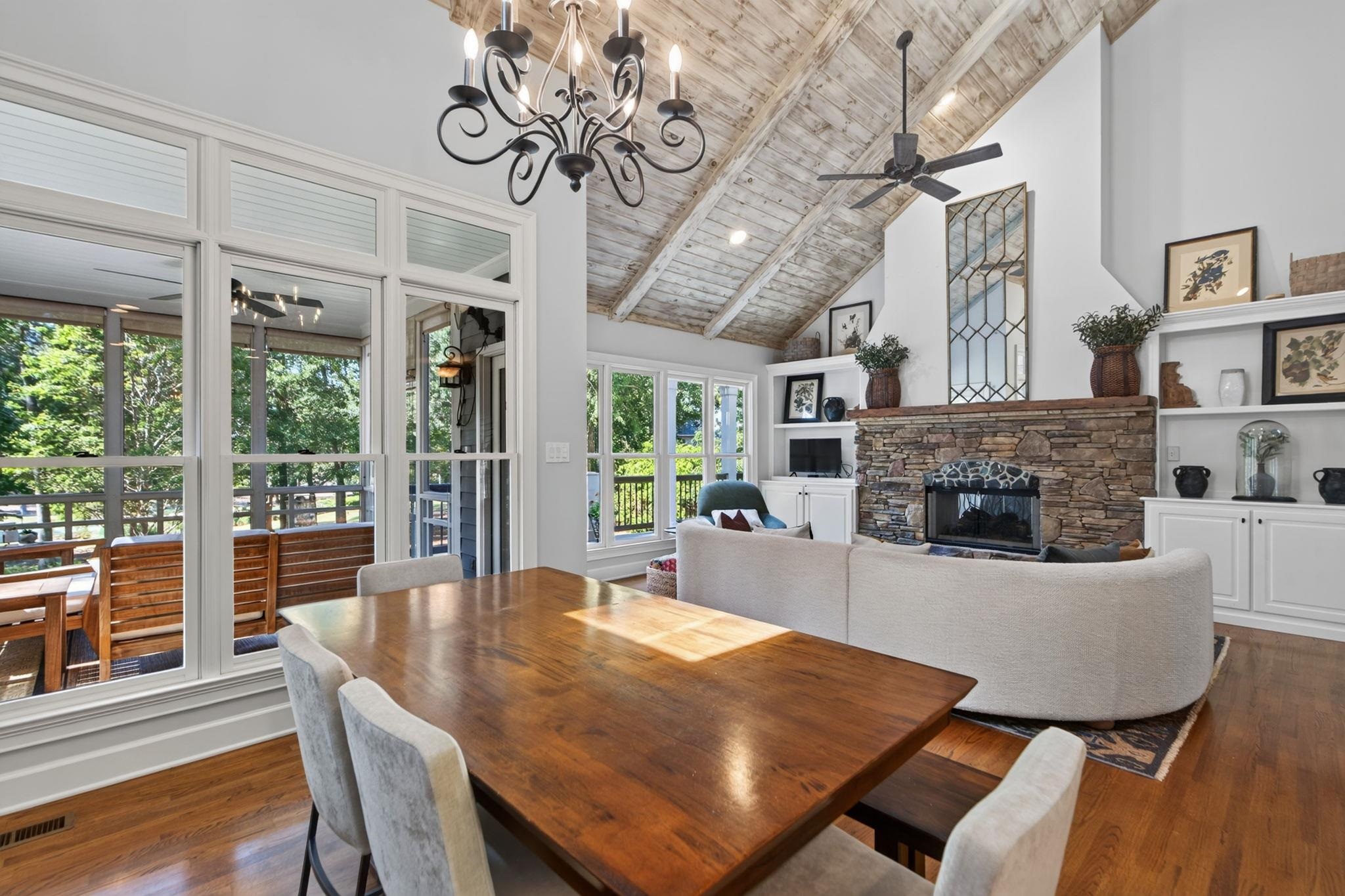 Living and dining room with a vaulted wood ceiling, stone fireplace, and windows overlooking a tree-lined deck.