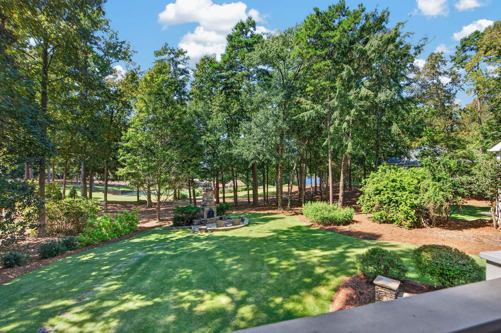 Wooded backyard with a broad lawn and stone outdoor fireplace beside a glimpse of water.