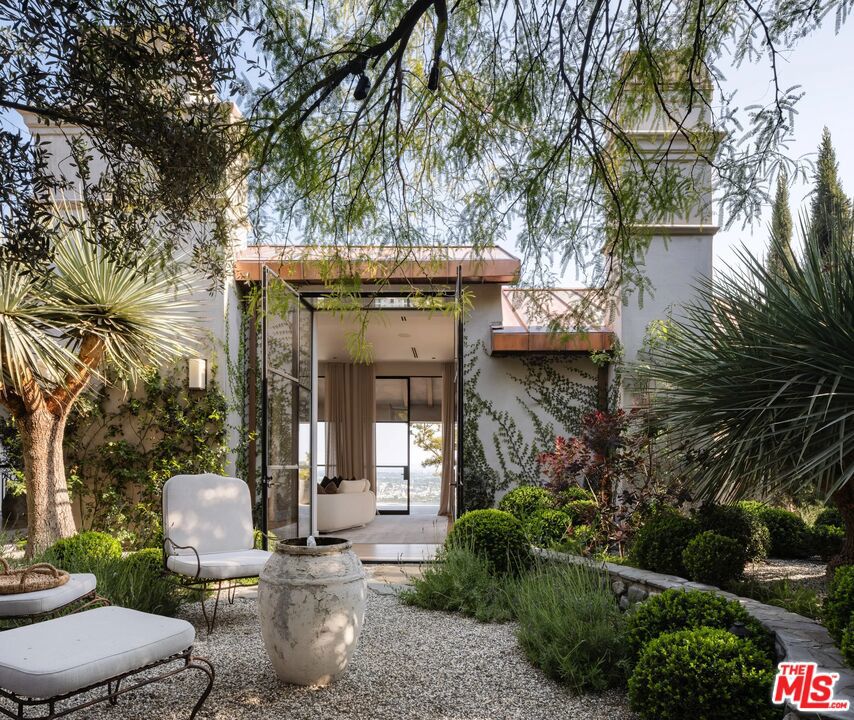 Modern home entrance framed by a leafy courtyard, with glass doors and outdoor lounge seating.