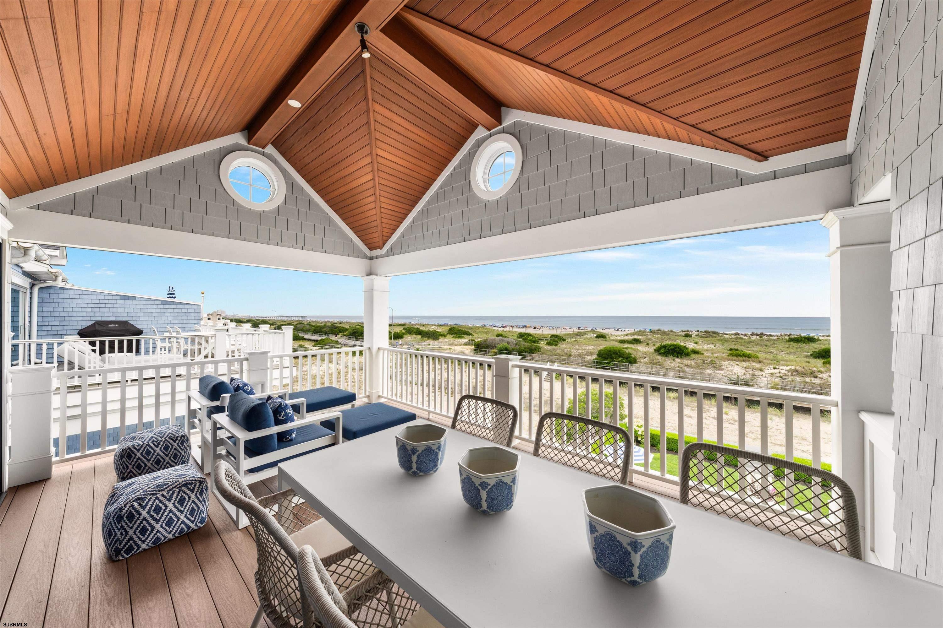 Covered oceanfront deck with dining and lounge seating beneath a vaulted wood ceiling, overlooking beach dunes.