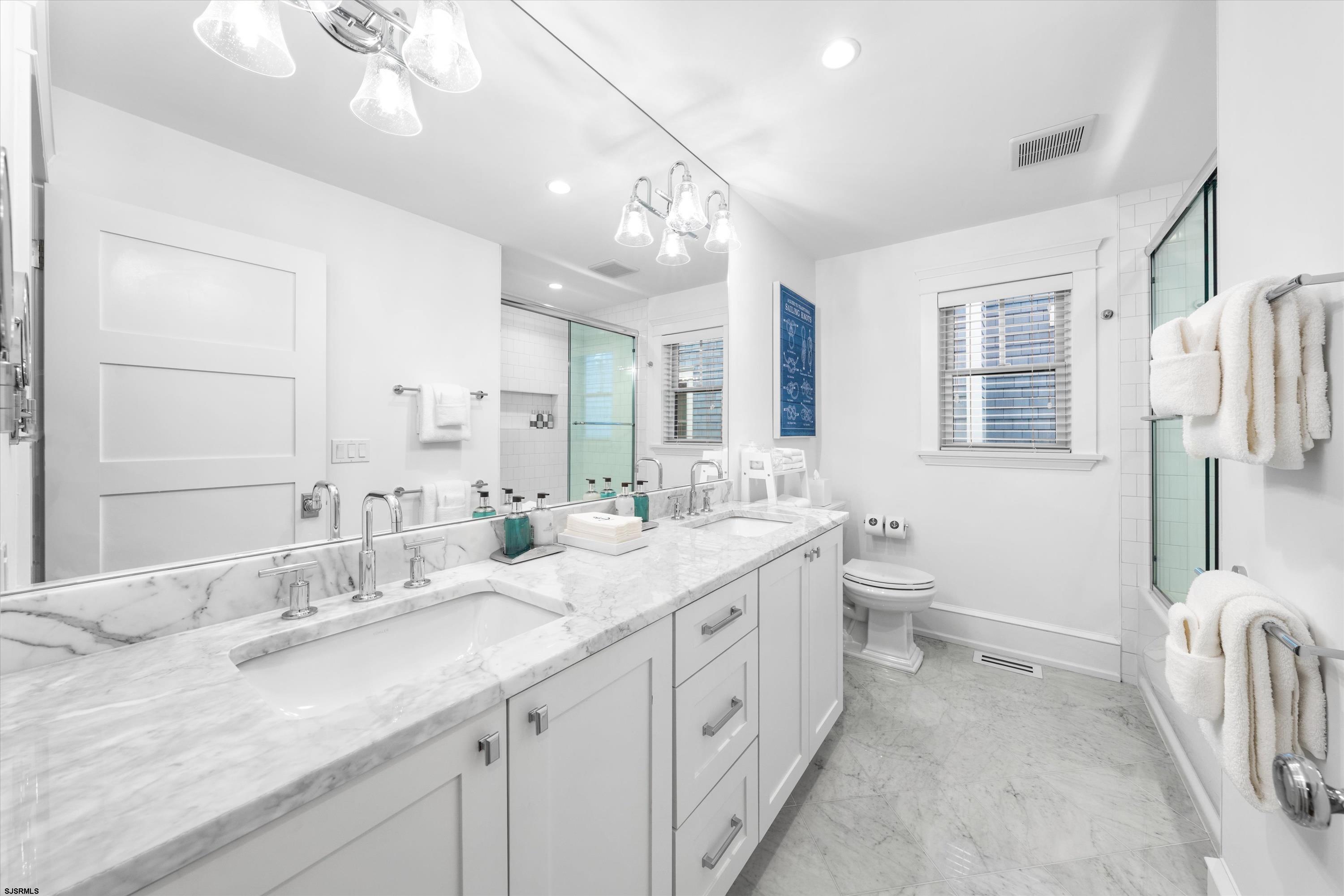 Bright bathroom with a marble double vanity, glass shower, and window with blinds.