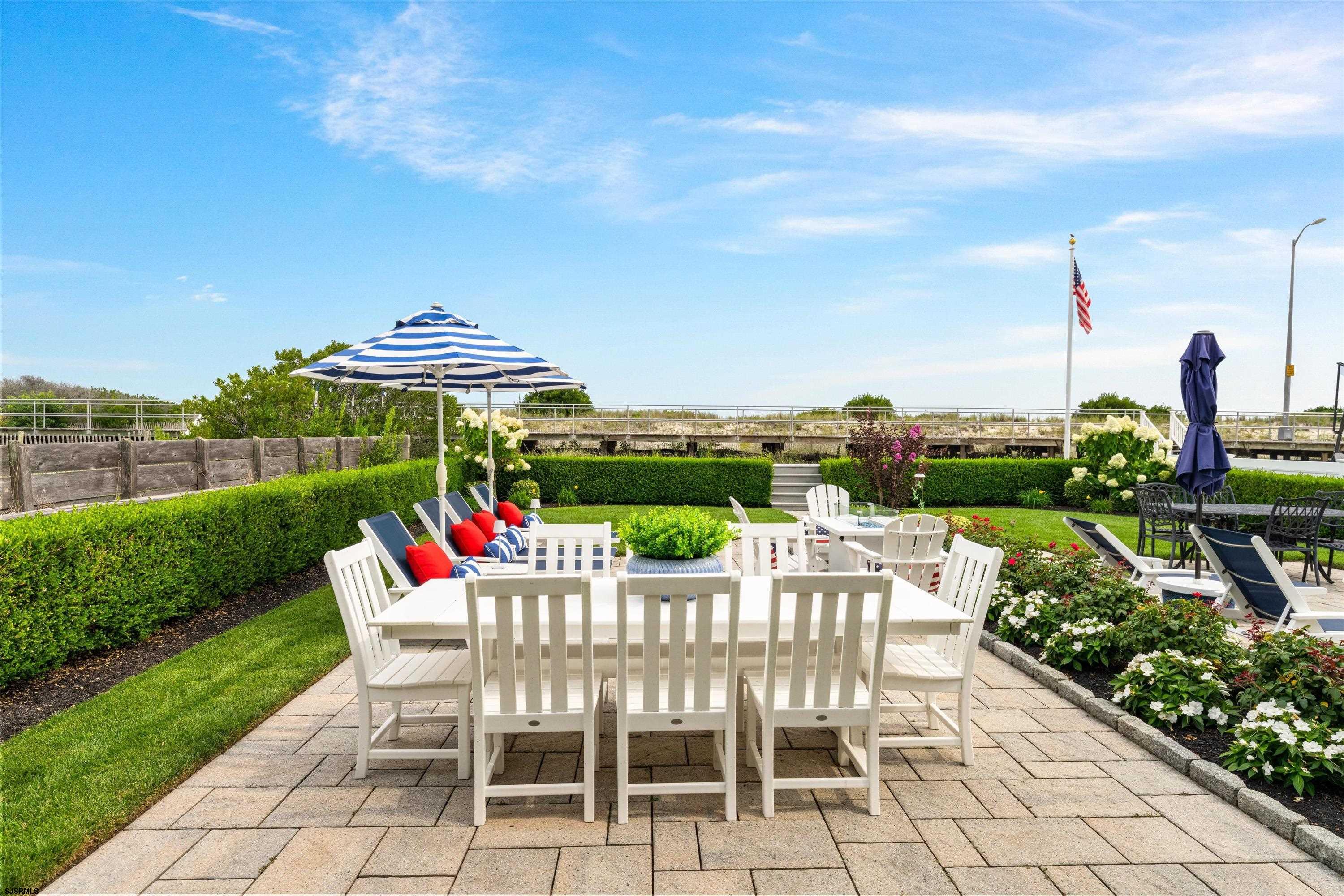 Outdoor patio with white dining furniture, a striped umbrella, lounge chairs, and landscaped flower beds.