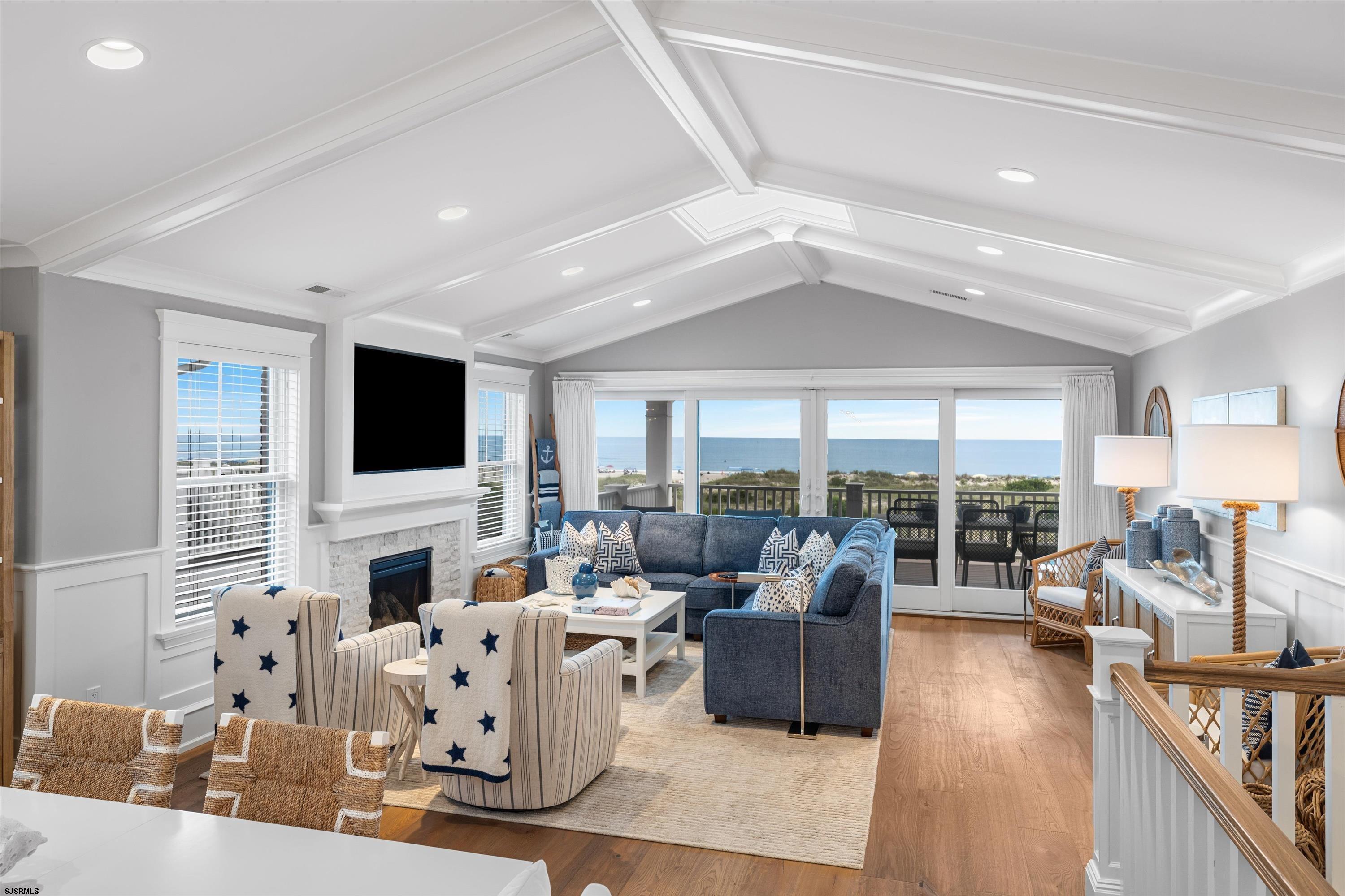 Coastal living room with a vaulted ceiling, blue-gray sectional, fireplace, and ocean view through sliding glass doors.