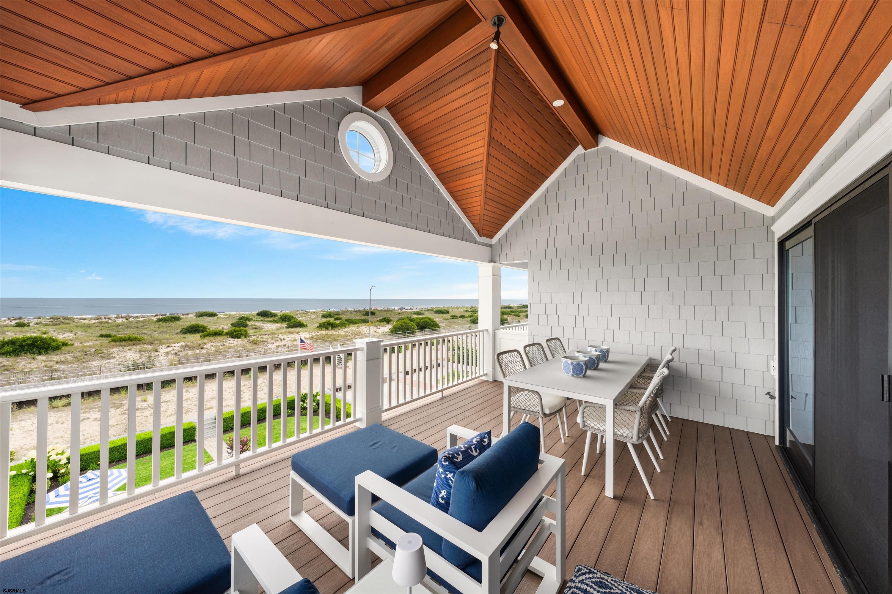 Covered deck with blue lounge seating and dining table overlooking dunes and the ocean.