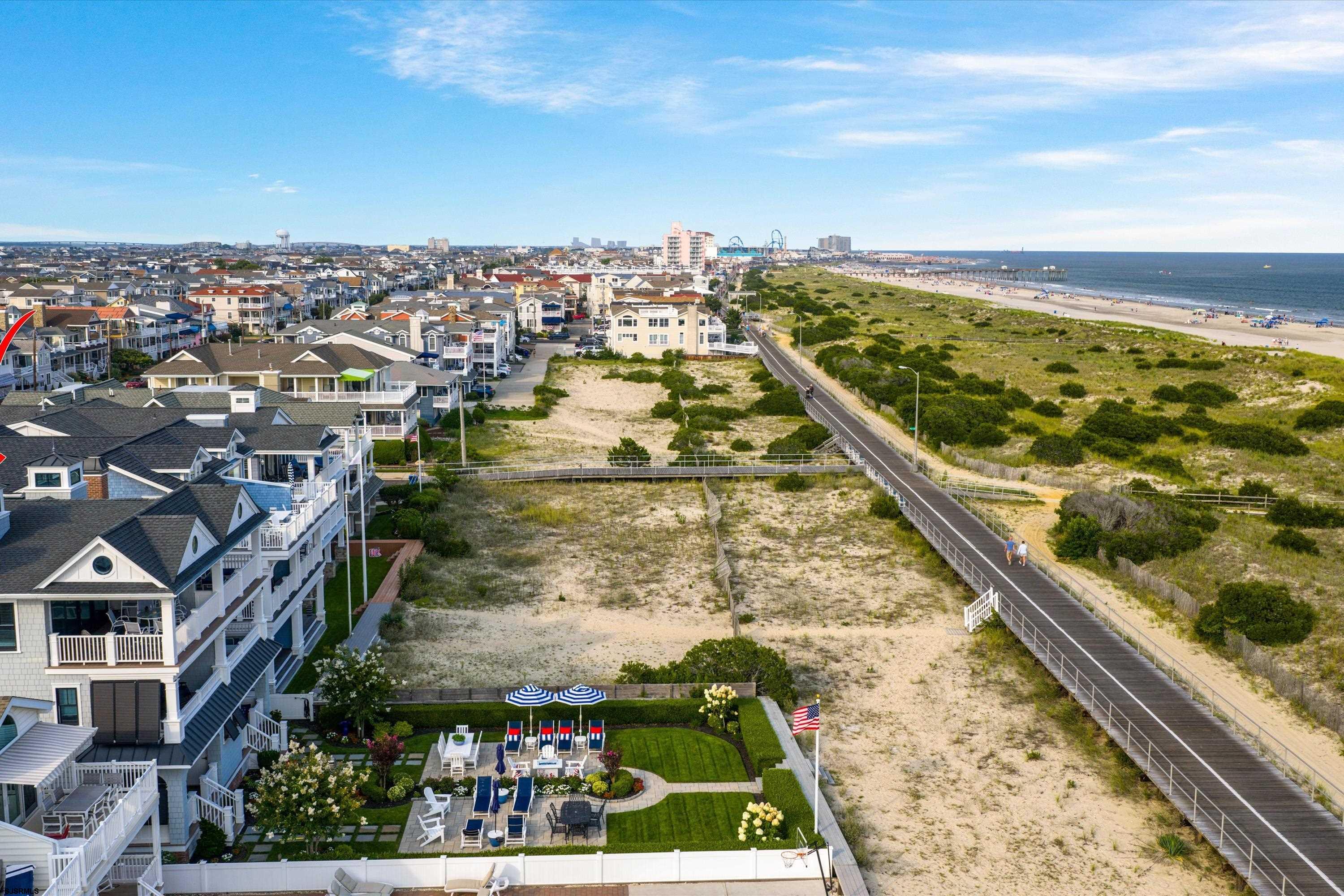 Aerial view of beachfront homes, a vacant sandy lot, dunes, boardwalk, and ocean.