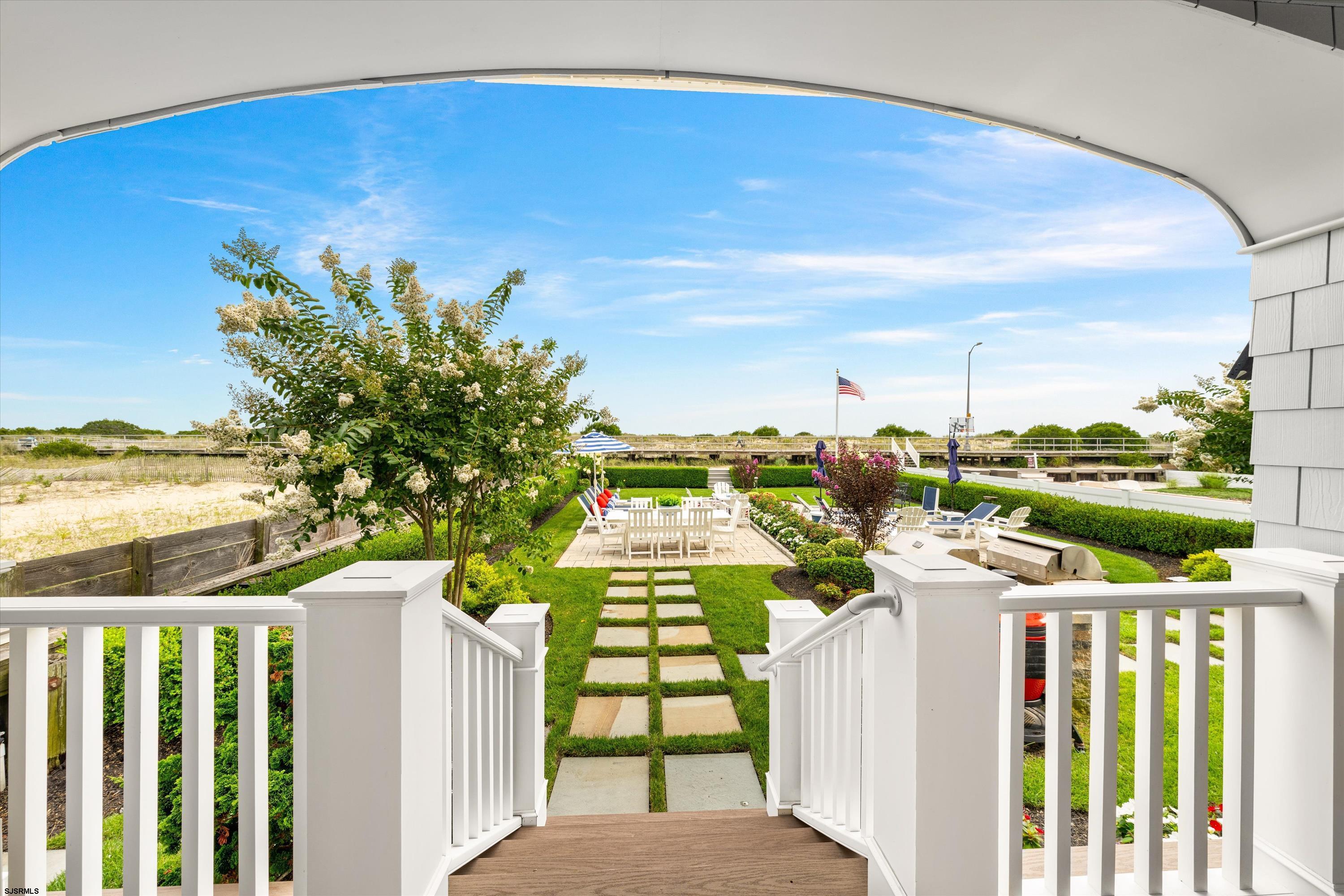 Landscaped garden patio with a stepping-stone path, white dining chairs, flowering trees, and a boardwalk beyond.