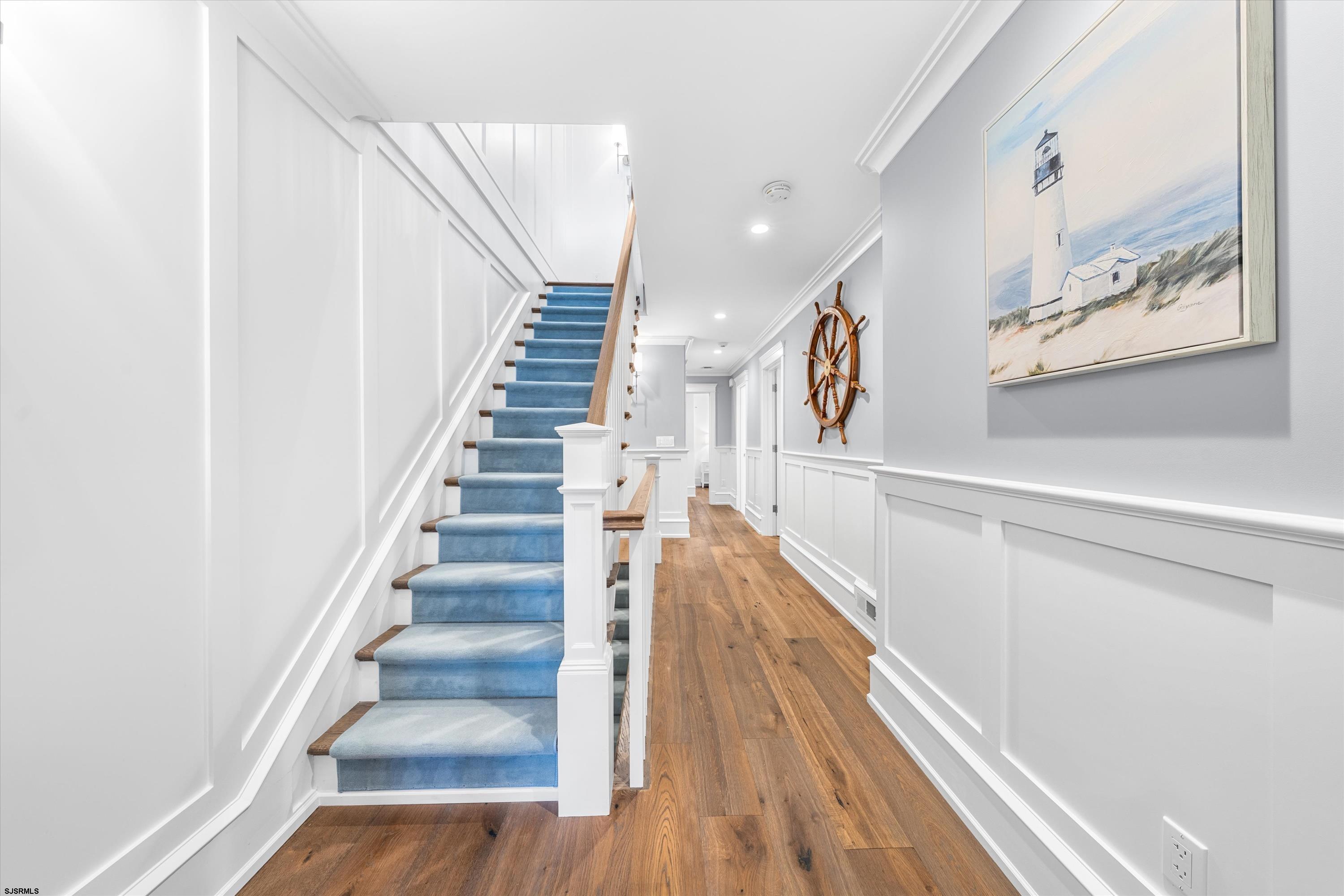 Hallway with blue-carpeted stairs, wood flooring, and coastal decor.