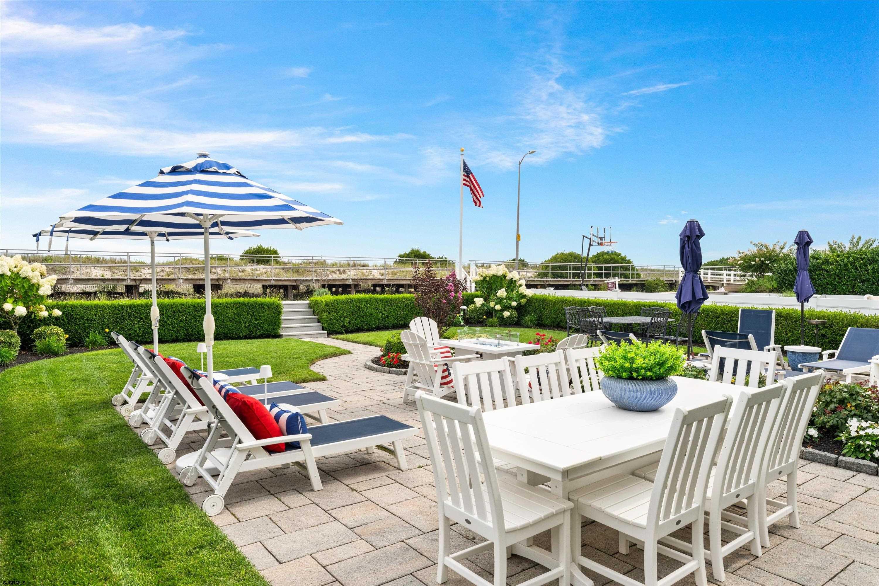 Outdoor patio with white dining furniture, lounge chairs, striped umbrellas, and landscaped flower beds.