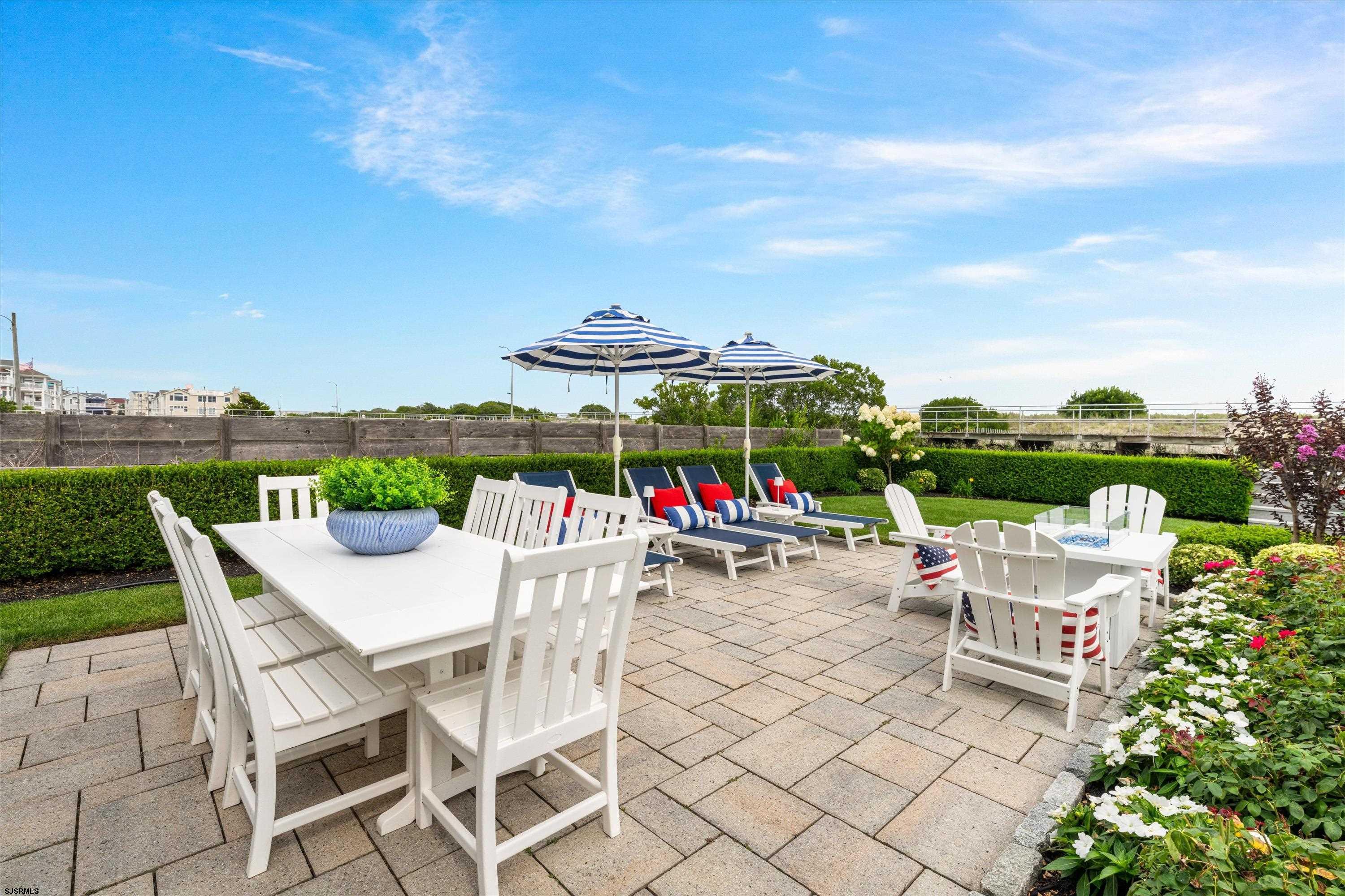 Outdoor patio with white dining furniture, striped umbrellas, and lounge chairs beside landscaped flower beds.