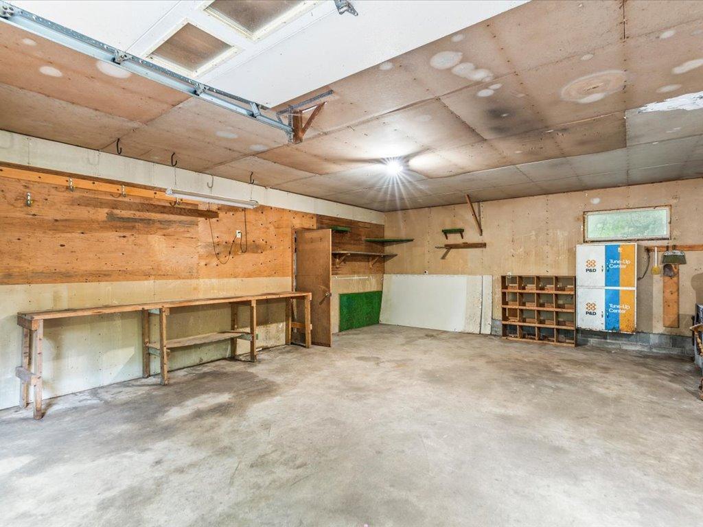 Garage workshop with a concrete floor, long workbench, shelving, and exposed ceiling panels.