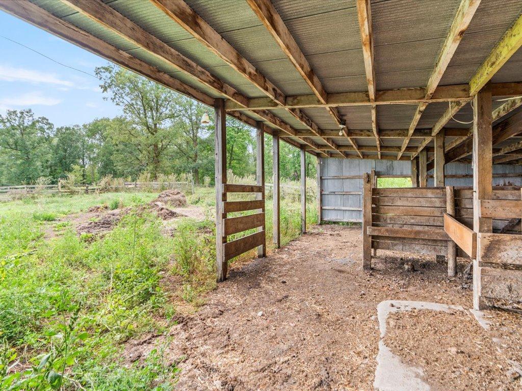 Open-sided wooden livestock shelter with stall partitions and a dirt floor beside a grassy yard.