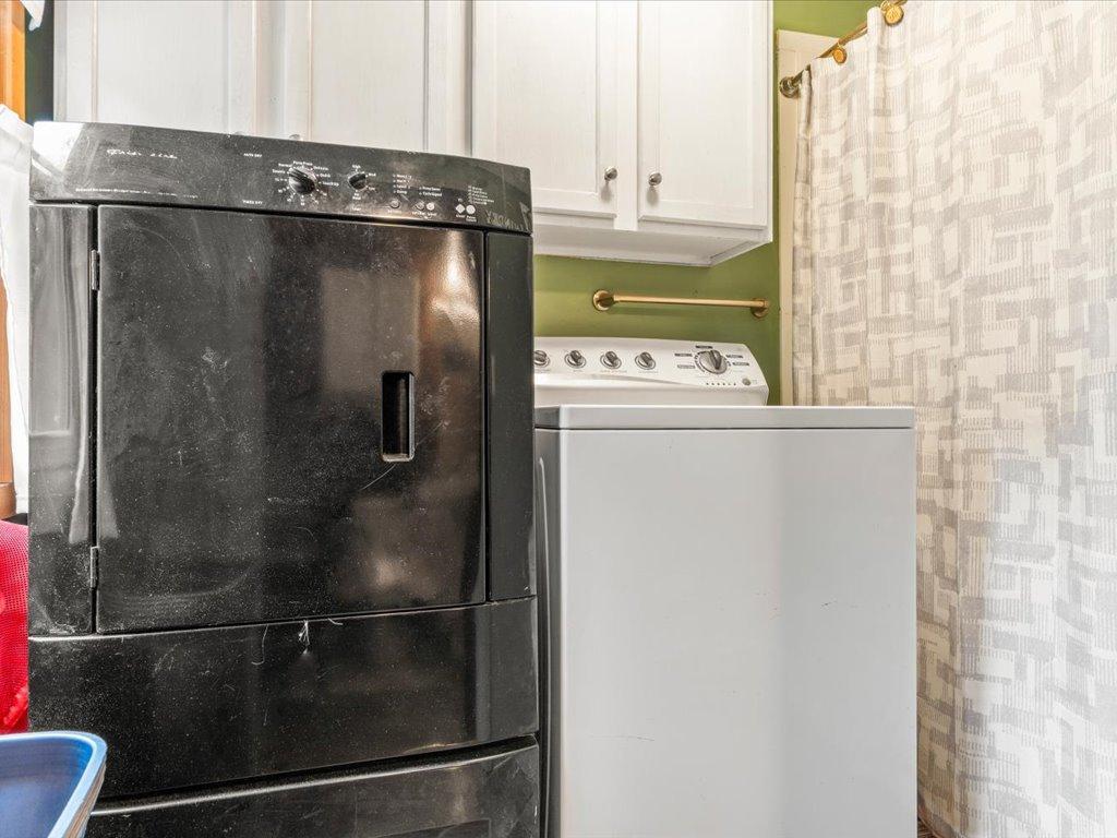 Laundry room with a black dryer, white washer, overhead cabinets, and patterned curtain.