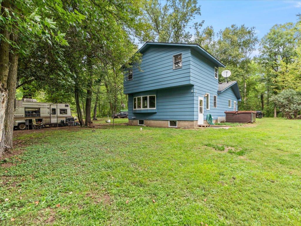 Blue two-story house in a wooded yard, with a travel trailer parked beneath the trees.