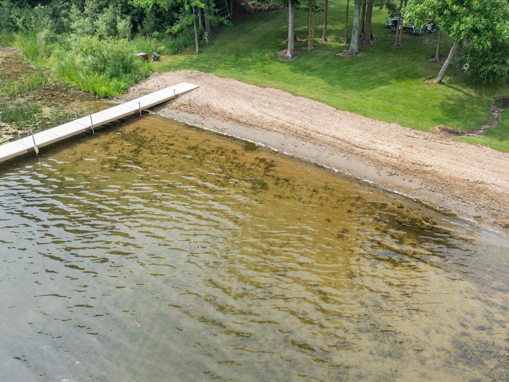 Sandy lakeshore with a long white dock, clear shallow water, and a wooded grassy yard.