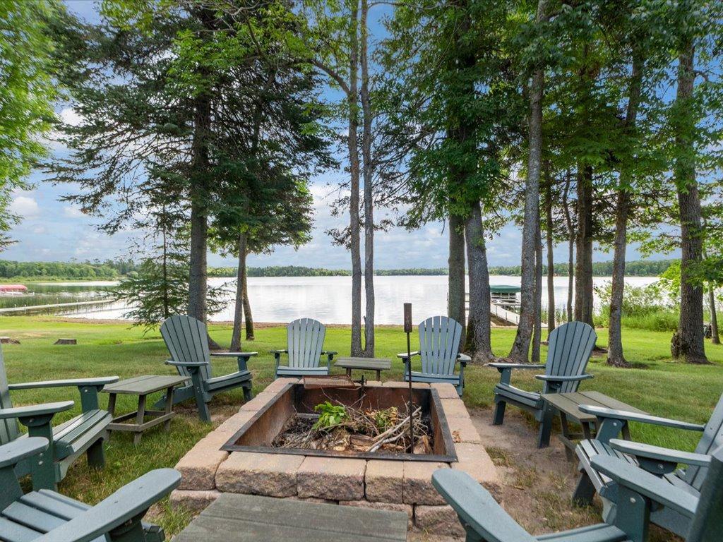 Lakeside patio with Adirondack chairs around a stone fire pit beneath mature trees.