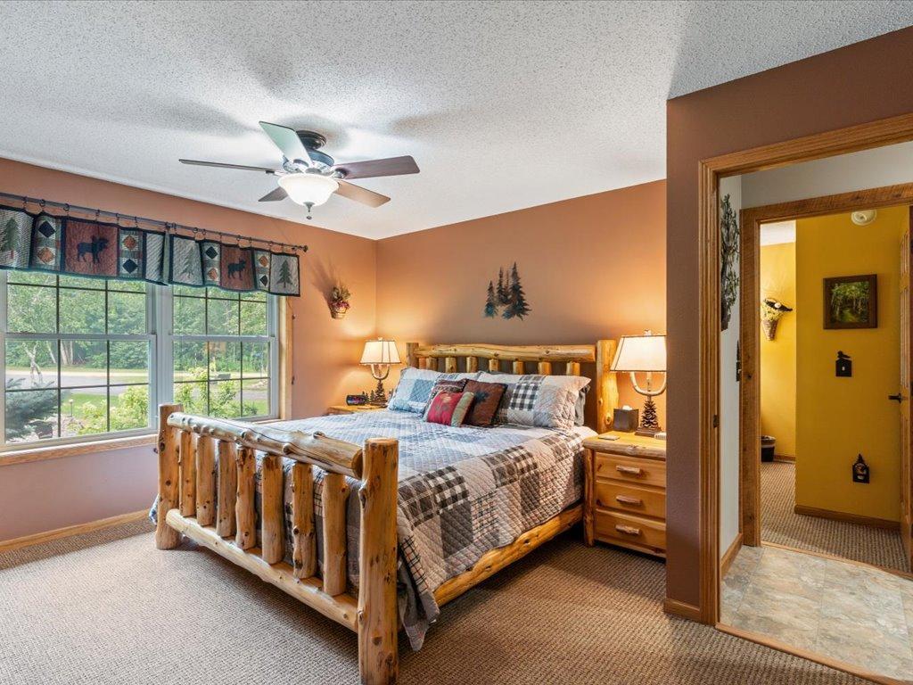Furnished bedroom with a rustic log bed, plaid bedding, ceiling fan, and large windows overlooking greenery.