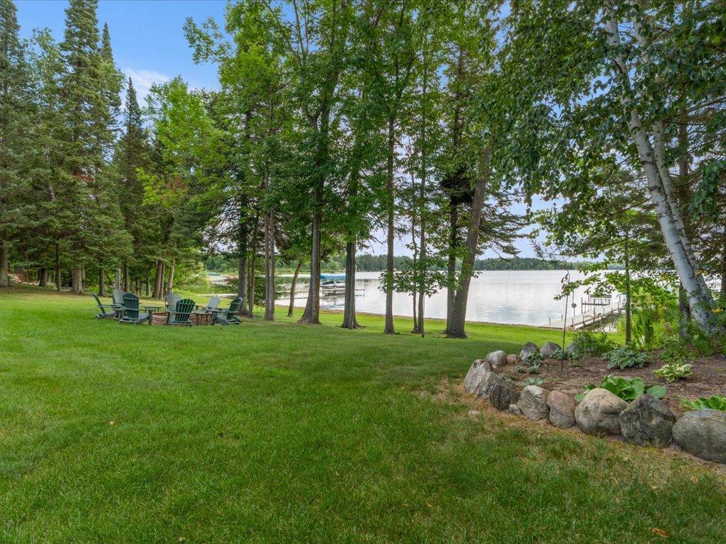 Lakeside yard with mature trees, Adirondack chairs, and a dock on the water.