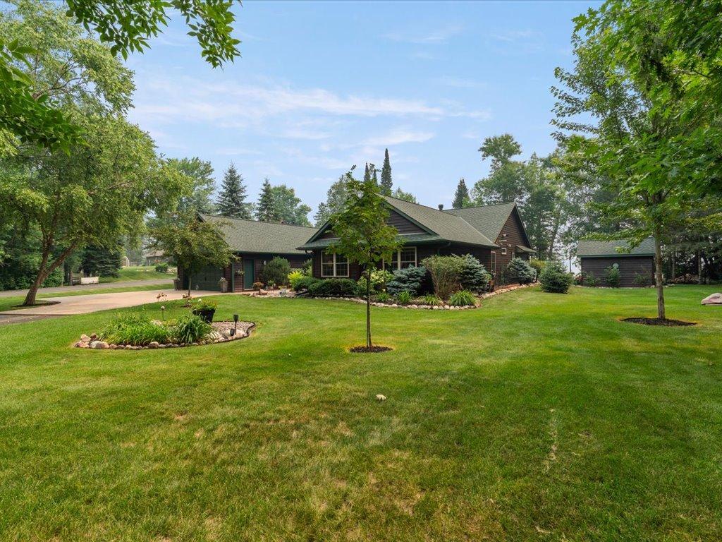 Single-story home behind a broad lawn, with mature trees, garden beds, and a detached garage.