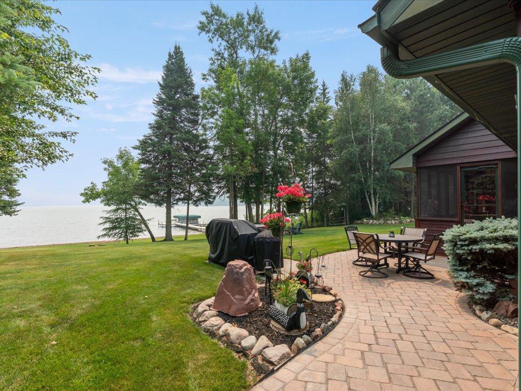 Lakeside yard with a brick patio, outdoor dining set, flower beds, and a dock beyond the lawn.
