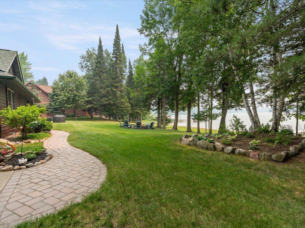 Lakeside yard with a curved paver walkway, mature trees, and chairs beside the water.