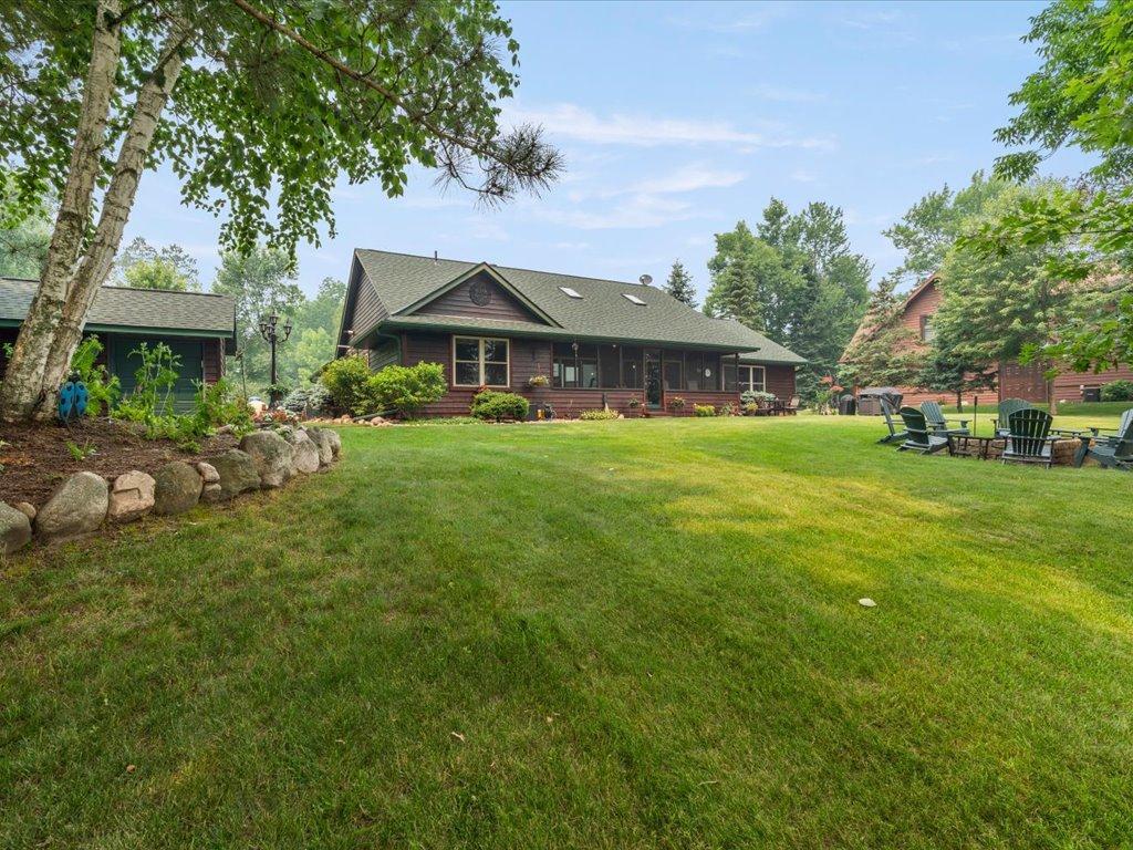 Single-story wood house with a covered porch, broad lawn, mature trees, and Adirondack chairs by a fire pit.