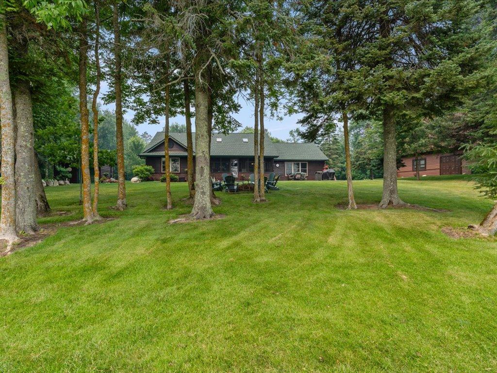 Wood-sided home beyond a broad lawn, framed by mature trees, with a separate outbuilding nearby.