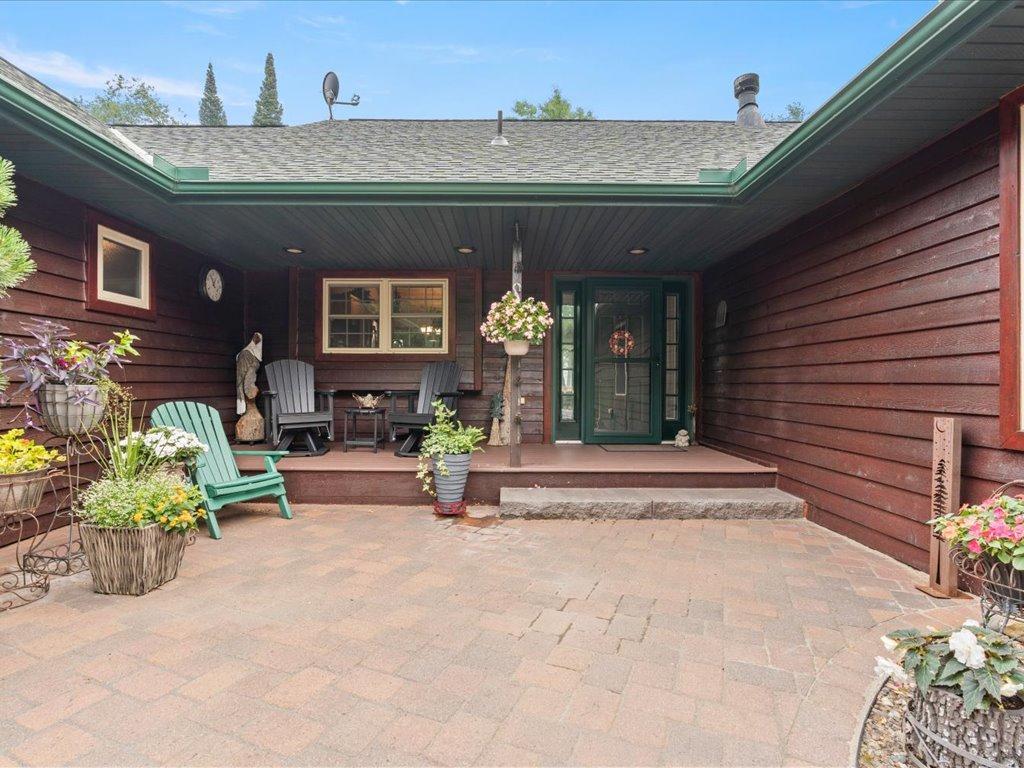 Covered entry patio with a green-trimmed glass door, paver flooring, Adirondack chairs, and potted flowers.