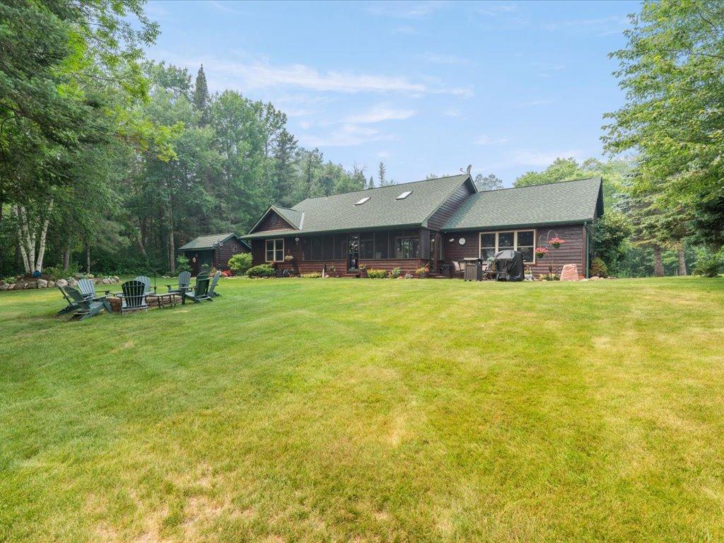 Single-story dark wood house with a broad lawn, wooded surroundings, and outdoor seating.