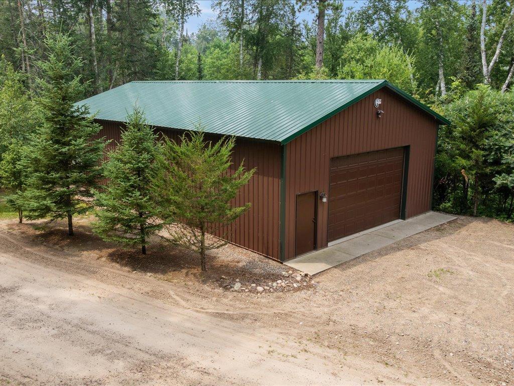 Brown detached garage with a green metal roof, wide overhead door, and wooded surroundings.