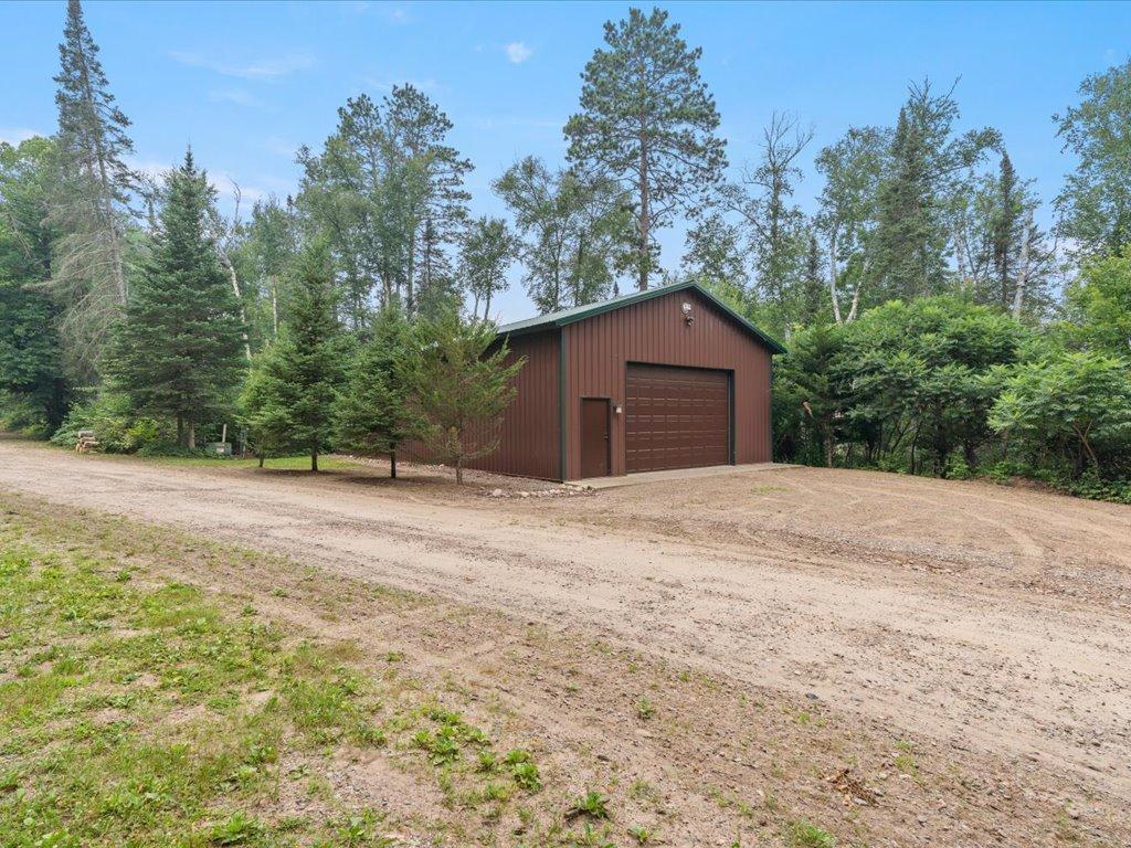 Brown detached garage with a wide overhead door beside a broad gravel driveway and wooded surroundings.