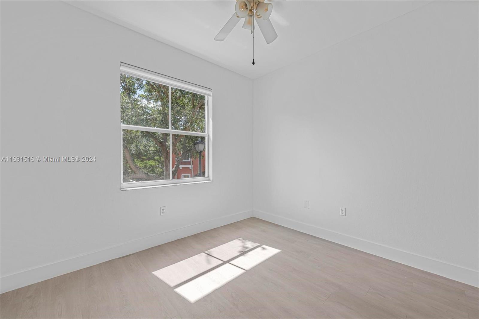 Empty room with light wood-look flooring, a ceiling fan, and sunlight streaming through a large window.